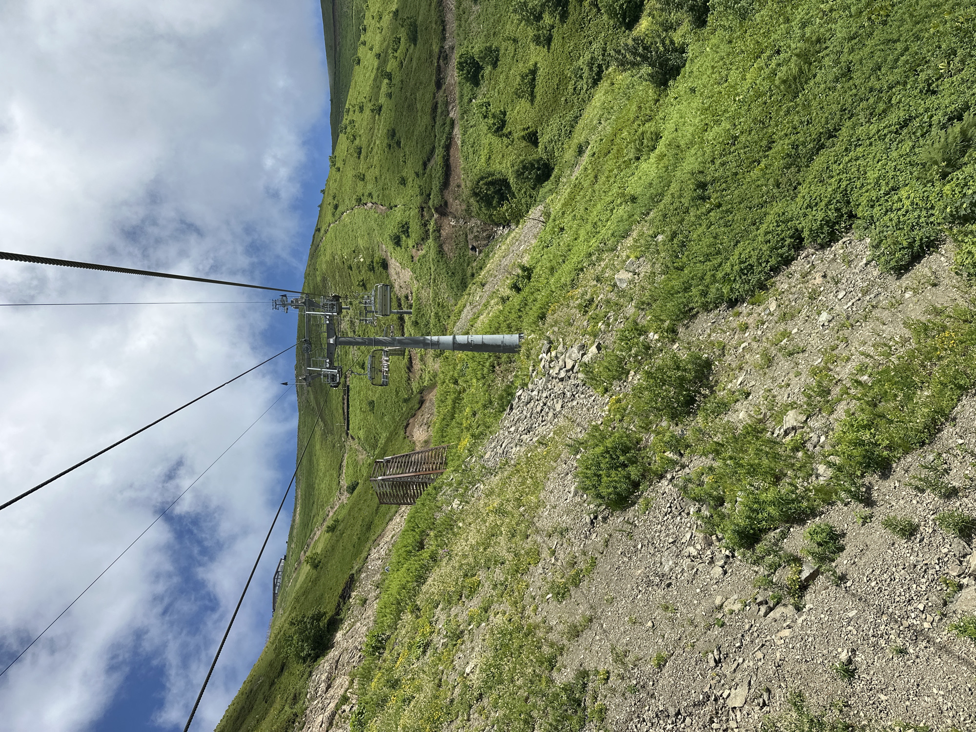 A ski lift ascends a lush green mountain under a partly cloudy sky. The ski lift's metal poles and cables stretch diagonally across the frame, with empty chairs hanging from the cables. The mountain is covered in thick green grass and dotted with small bushes and wildflowers. A rocky, dirt path winds its way up the slope to the right of the ski lift. In the distance, a building with a dark roof is visible on the mountaintop. A wooden structure, possibly a retaining wall or a part of the ski lift infrastructure, is situated on the left side of the mountain. The terrain is steep and uneven, with a mix of rocky ground and dense vegetation. The overall scene depicts a scenic mountain landscape, likely during the summer months.