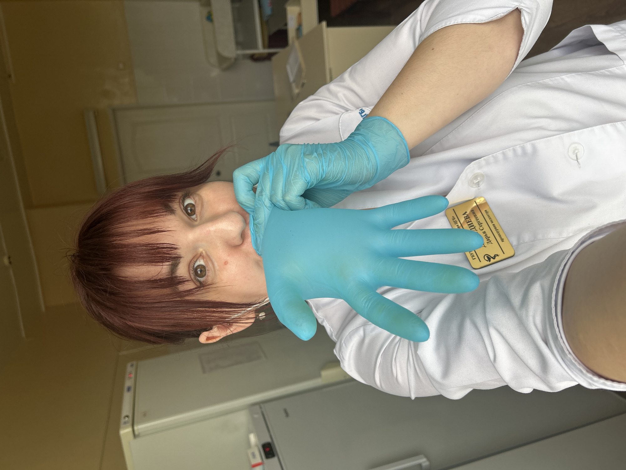 A fair-skinned woman with brown hair and bangs is wearing a white lab coat and blue latex gloves. She is in a room with white walls and appliances, possibly a laboratory or medical facility in Фоки, Russia. She is pulling one glove off, using the other gloved hand to grip the first glove at the fingertips. The image is oriented vertically.