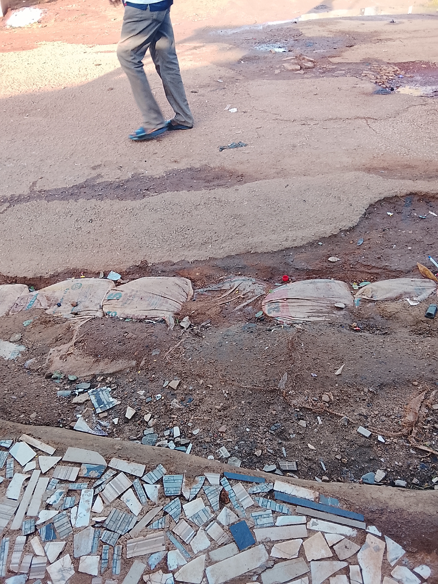 The image captures a street scene in Kabong, Nigeria, characterized by disrepair and informal construction. In the upper portion, a person is walking on a paved surface, their legs clad in grey trousers and dark blue flip-flops. The ground is uneven, with patches of water and rubble. The lower part of the image reveals a construction area, possibly for drainage or road repair. Several large sacks, filled with what appears to be sand or cement, are laid out in a trench, acting as barriers. The surrounding earth is disturbed, with loose soil and debris. Scattered across the ground are numerous broken pieces of tile, some with intricate patterns in blue and white, suggesting remnants of a mosaic or decorative surface. The overall impression is one of ongoing, perhaps temporary, work in a less-maintained urban environment.