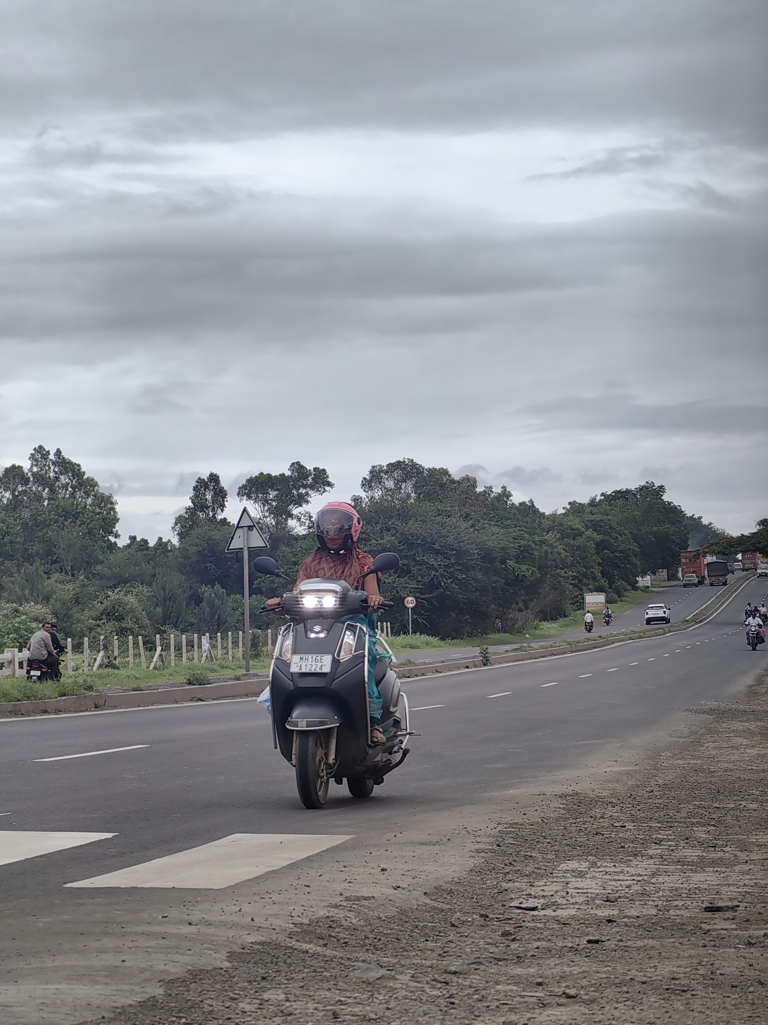 The image shows a cloudy day on a road in the vicinity of Chas, India. In the foreground, a person wearing a helmet and a patterned garment is riding a scooter with its headlights on. The scooter has a license plate that reads "MH16E A1224". To the left of the scooter, two men are standing near another scooter, with a fence and lush green trees in the background. The road stretches ahead, with several vehicles visible in the distance, including trucks, cars, and other motorcycles. The sky is overcast with heavy grey clouds. The road surface appears to be asphalt, with some gravel and patches of damage. White lines mark the lanes on the road, and there are pedestrian crossings visible in the foreground. The overall scene depicts a typical highway environment with traffic and roadside vegetation.