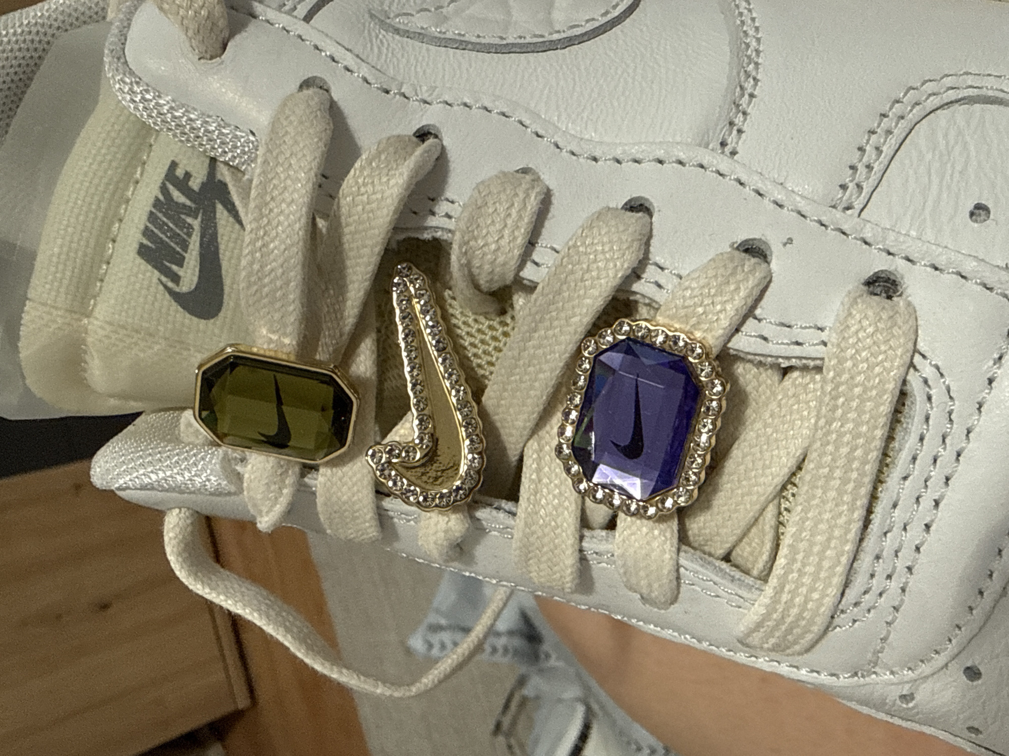 A close-up shot captures the top section of a white Nike sneaker, adorned with several decorative elements. The shoe itself appears to be made of white leather or synthetic material, featuring subtle stitching details along the laces. Off-white, flat woven shoelaces are threaded through the eyelets.
Three distinct, jewel-like lace charms (dubraes) are prominently displayed on the laces. The leftmost charm is a rectangular, olive green facet-cut gem set in a gold-colored frame, with a small black Nike swoosh logo embedded within it. The middle charm is a stylized gold-colored "J" shape, encrusted with numerous small clear rhinestones or crystals. The rightmost charm is an octagonal or cushion-cut deep purple facet-cut gem, also framed in gold-colored metal and surrounded by small clear rhinestones, featuring another black Nike swoosh logo inside.
Visible on the shoe's tongue, behind the laces, is the iconic "NIKE" text logo in black. The background of the image is softly blurred, showing hints of a warm-toned, possibly wooden, surface. The overall impression is a detailed showcase of personalized footwear accessories. The image does not contain any visual cues pertaining to the specified location of Hainburg an der Donau, Austria.