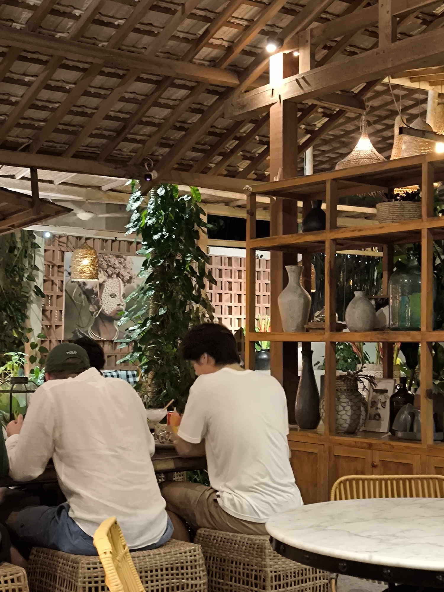 This media file captures an indoor or semi-outdoor restaurant or cafe scene in North Kuta, Indonesia, illuminated by warm artificial light, indicating it is evening. Two men are seated at a table, viewed from behind. The man on the left wears a white long-sleeved shirt, blue shorts, and a green baseball cap with the brand name "POLO" clearly visible. He sits on a woven stool. The second man, on the right, wears a white t-shirt and tan shorts, also seated on a similar woven stool.
The setting is characterized by a rustic and natural aesthetic. An exposed wooden beam and rafter ceiling dominates the upper portion of the frame, with visible roofing material above the beams. Lush green plants and vines are abundant, integrated into the decor and growing against walls and pillars. Various light sources, including woven pendant lamps, exposed bulbs, and spotlights, create a warm and inviting atmosphere.
To the right, a tall wooden shelving unit acts as a decorative partition, filled with an array of vases in shades of grey, black, and clear glass, along with woven baskets. A round, white marble-top table with a dark base is partially visible in the lower right foreground. In the mid-ground, a large piece of art depicting a face with intricate patterns is mounted on a wall featuring decorative open brickwork. A small white ceiling fan and a security camera are noticeable fixtures overhead. The space utilizes natural materials like wood and woven textures, contributing to a relaxed, bohemian, and tropical ambiance.