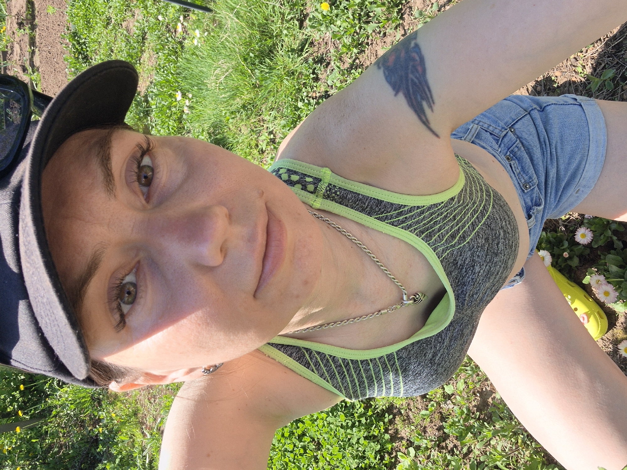 A woman wearing a black baseball cap, a gray sports bra with green trim, and denim shorts is lying on a patch of grass with dandelions and daisies. She is wearing a silver chain necklace and has a tattoo on her upper left arm. One bright yellow shoe is visible near the daisies. The location is likely in a grassy area in Chaikovsky, Russia, given the context.