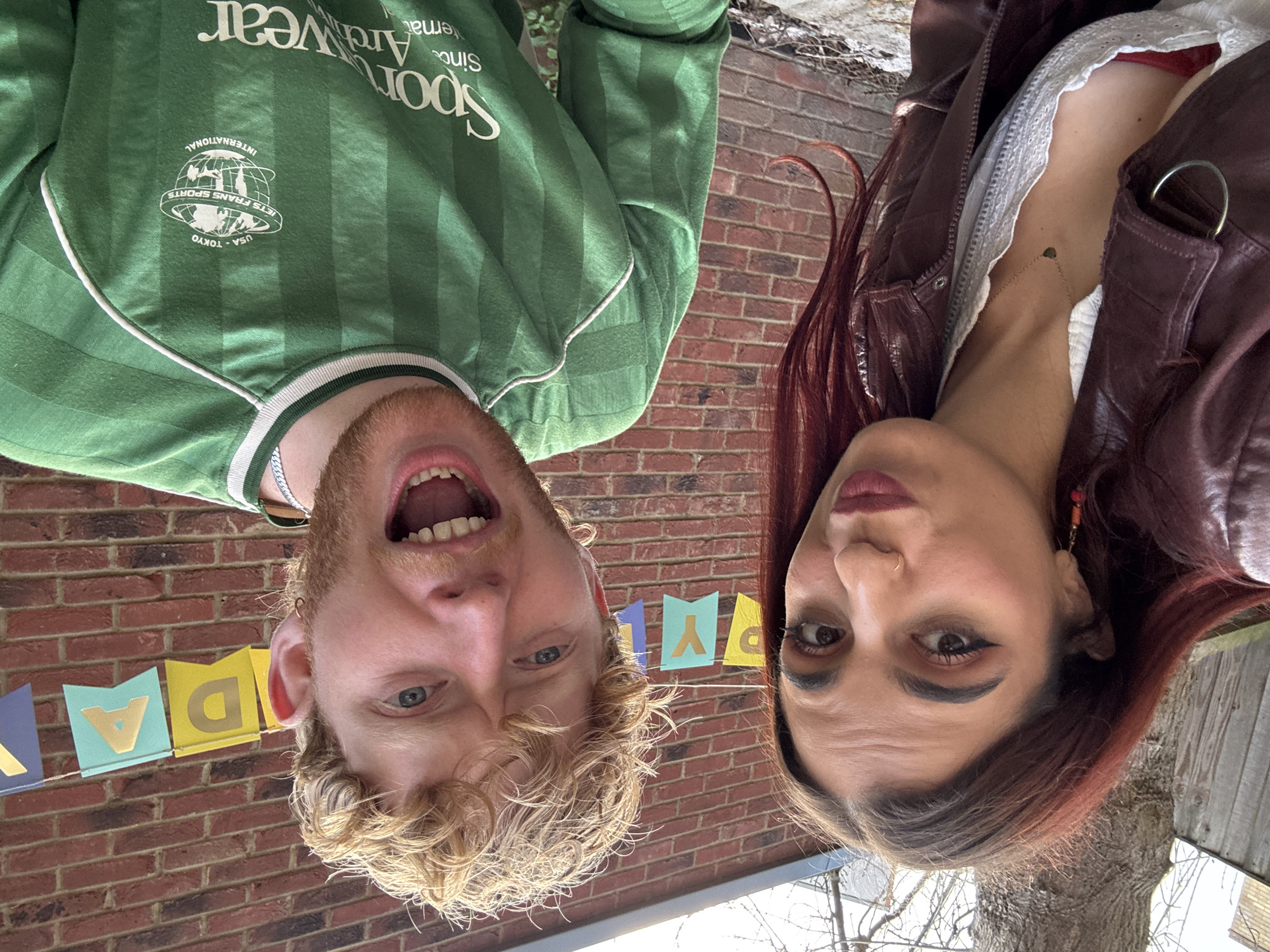 Two individuals pose closely for a selfie. A man with blond, curly hair and blue eyes is on the left, wearing a green retro soccer jersey with the text "Sportswear" on it, and a silver chain. He has his mouth open in a wide smile. A woman with dark hair and makeup is on the right, wearing a burgundy leather jacket and a delicate gold necklace. Both are upside down relative to the backdrop. Behind them is a red brick wall with a colorful "Happy Birthday" banner strung across. Further in the background, there are glimpses of trees and possibly a fence, suggesting an outdoor setting. There are no features in the image that suggest the city of London, United Kingdom.