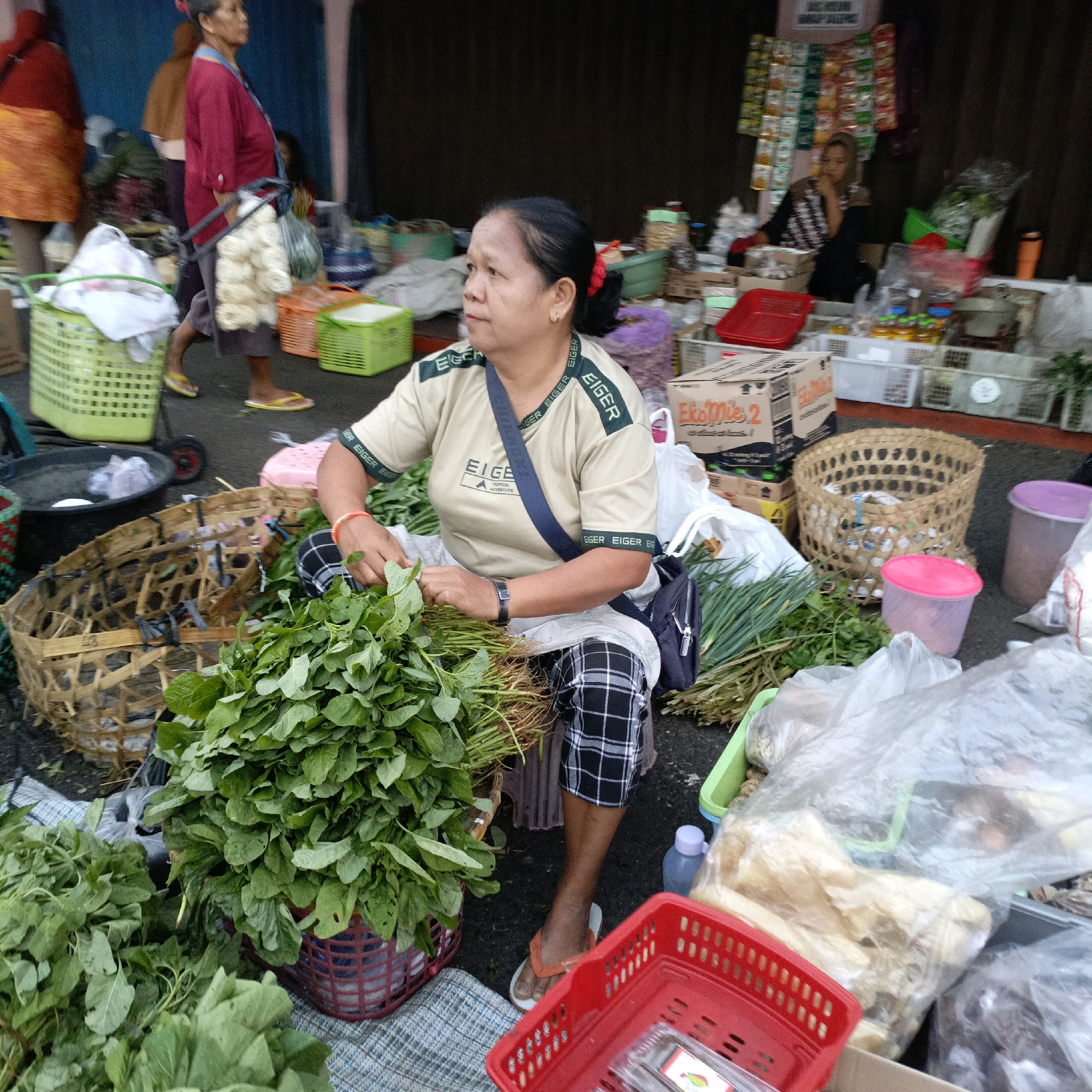 This is a medium-shot, eye-level photograph taken at a street market in Salatiga, Indonesia. The foreground is dominated by a vendor, a woman with dark hair pulled back and wearing a light-colored t-shirt with "EIGER" branding and checkered pants. She is seated and appears to be sorting or arranging fresh green vegetables, specifically leafy greens and what look like green onions, from a woven basket. Her hands are actively engaged with the produce.
Around her, the environment is cluttered with various market items. To her left, a large woven basket sits empty. In front of her, a red plastic basket contains more produce. Other baskets and containers, some green and some red, are visible, along with plastic bags containing goods, and stacked cardboard boxes. Two pink plastic tubs are placed near the woven basket. In the midground, other market stalls and vendors are partially visible, including a woman in a maroon shirt carrying a bundle of items and another woman in a headscarf seated behind a stall with packaged goods.
The ground is a dark, paved surface. The overall lighting suggests it is daytime, possibly morning or late afternoon, with diffused natural light. The scene captures the bustling atmosphere of a local Indonesian market.