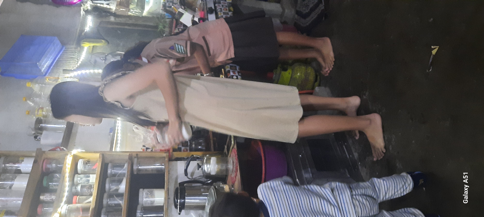 The image shows an indoor scene, likely a kitchen or pantry, with a shelving unit filled with jars and containers on the left. A young woman in a beige dress is standing in the center, holding a glass jar. To her right, another person, possibly a child, is wearing a brown and pink uniform with stripes on the sleeve. A green gas cylinder is visible behind them, and the floor appears to be damp or wet. The overall lighting is dim, with some artificial light sources creating a warm glow. The location is identified as Jatitengah, Indonesia.