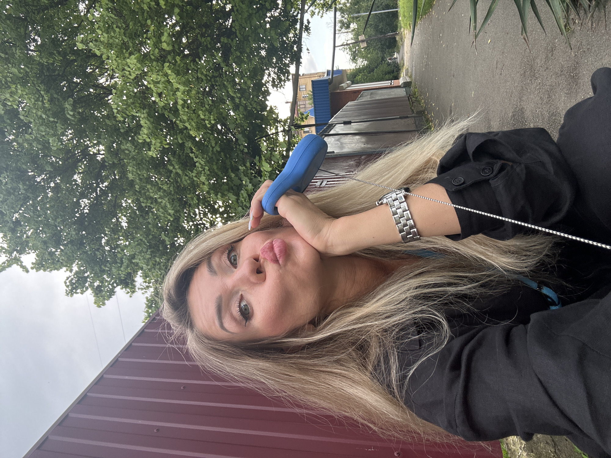 A blonde woman with light green eyes and heavy makeup is taking a selfie outdoors. She is wearing a black jacket over a blue top and a silver watch. She is puckering her lips and resting her hand on her chin. A blue object, possibly a dog leash handle, is in her right hand. In the background, there is a red metal fence, green trees, and a paved path leading past buildings. The location is identified as Armavir, Russia.