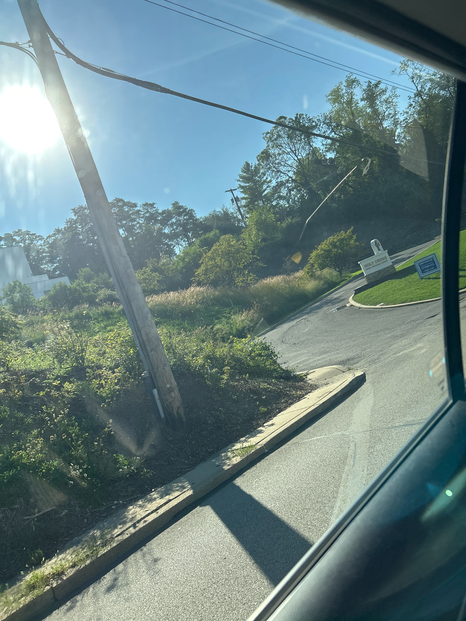 The image shows a view from inside a vehicle looking out at a street and a grassy incline. A wooden utility pole stands on the left side of the road, with power lines extending across the frame. The sun is bright and high in the sky, casting shadows and causing lens flare. On the right side of the road, there is a sign for "HOLY CROSS EVANGELICAL LUTHERAN CHURCH" with a stone base. The road curves to the right, leading towards the church entrance. Trees and vegetation cover the surrounding landscape. The scene appears to be a residential or suburban area. The specific location in Mt. Lebanon, United States of America, is not discernible from the image itself.
