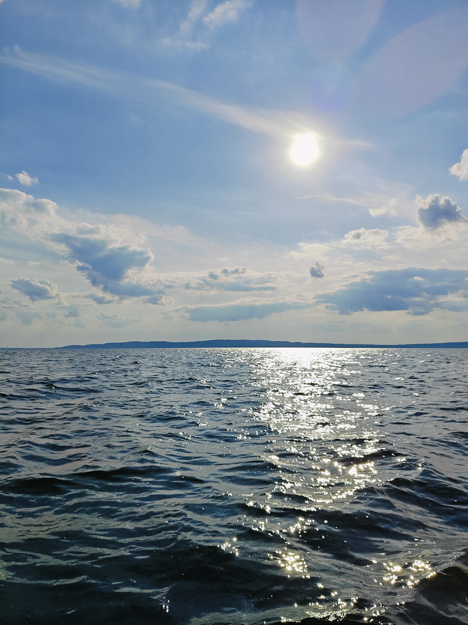 The image depicts a wide expanse of dark blue water with gentle waves, reflecting the bright sun and scattered white clouds in a clear blue sky. The sun is positioned high in the sky, casting a shimmering path of light across the water's surface. A distant, dark, and hilly shoreline is visible on the horizon. The scene is a natural landscape, likely a body of water like a lake or river, with no man-made structures or individuals immediately apparent. This scene could be located along the Kama River in Chaykovsky, Russia, known for its picturesque waterfront.