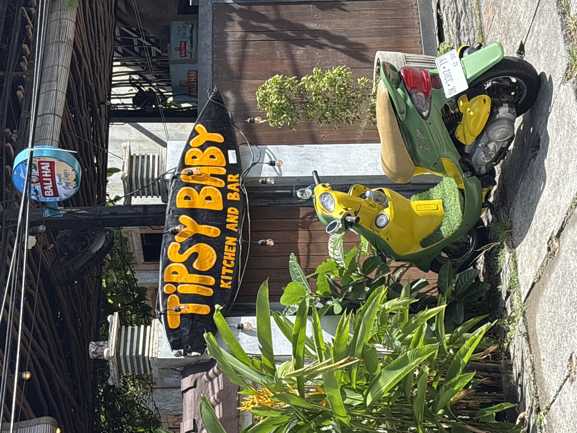 The image depicts a street scene in North Kuta, Indonesia. A sign for "TIPSY BABY KITCHEN AND BAR" is prominently displayed. A green and yellow scooter is parked nearby. The scooter's license plate reads "DK 3312 IV". The date on the plate is "12 29." Signage for "BALI HAI" beer is also visible. Lush vegetation surrounds the sign and scooter.