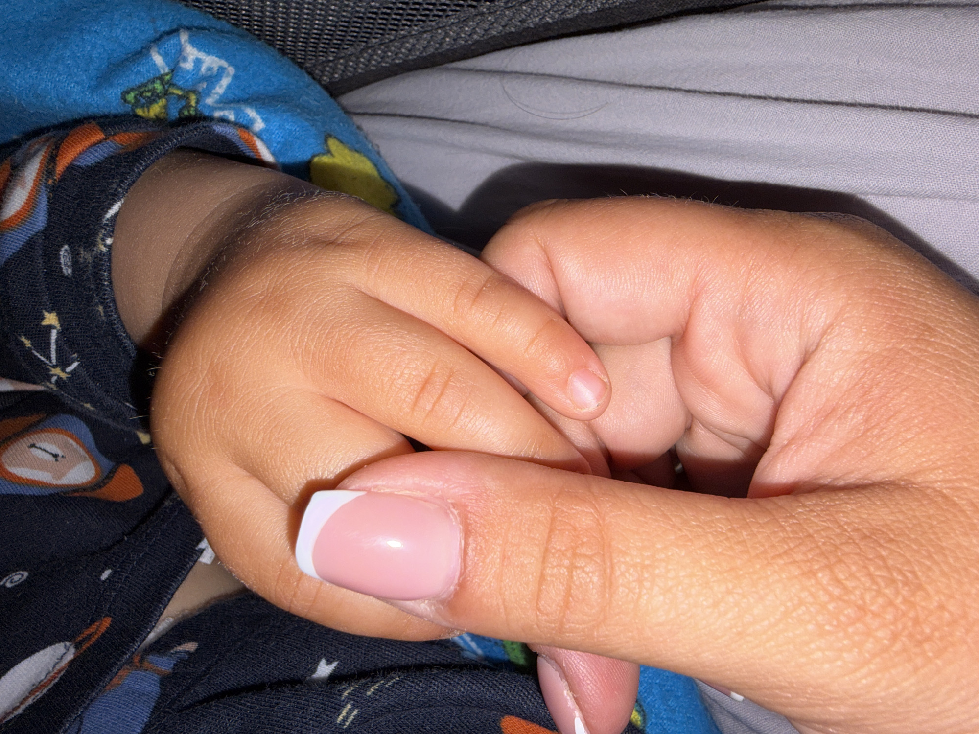 An intimate close-up shows an adult's hand gently holding a child's hand, conveying a sense of warmth and protection. The child's small, plump hand is clasped within the adult's fingers. The adult's fingernail, visible in the foreground, features a pink base with a white French tip.
The child is wearing a patterned blue garment, possibly pajamas, with various designs including what appears to be a green character or frog on the sleeve near the top, and other abstract or cartoon-like patterns on the darker blue fabric covering the lower arm. Part of a dark, mesh-like material, possibly from a crib, stroller, or playpen, is visible in the upper left corner and background, suggesting an indoor setting. Behind the hands, a light grey fabric, likely bedding or a blanket, is visible.
The lighting is soft and somewhat dim, casting subtle shadows and hinting at an evening or nighttime scene. The skin tones of both hands are warm, suggesting a tan complexion. No text is visible in the image. The scene, located in Svetovrachene, Bulgaria, evokes feelings of tenderness and care between the individuals.