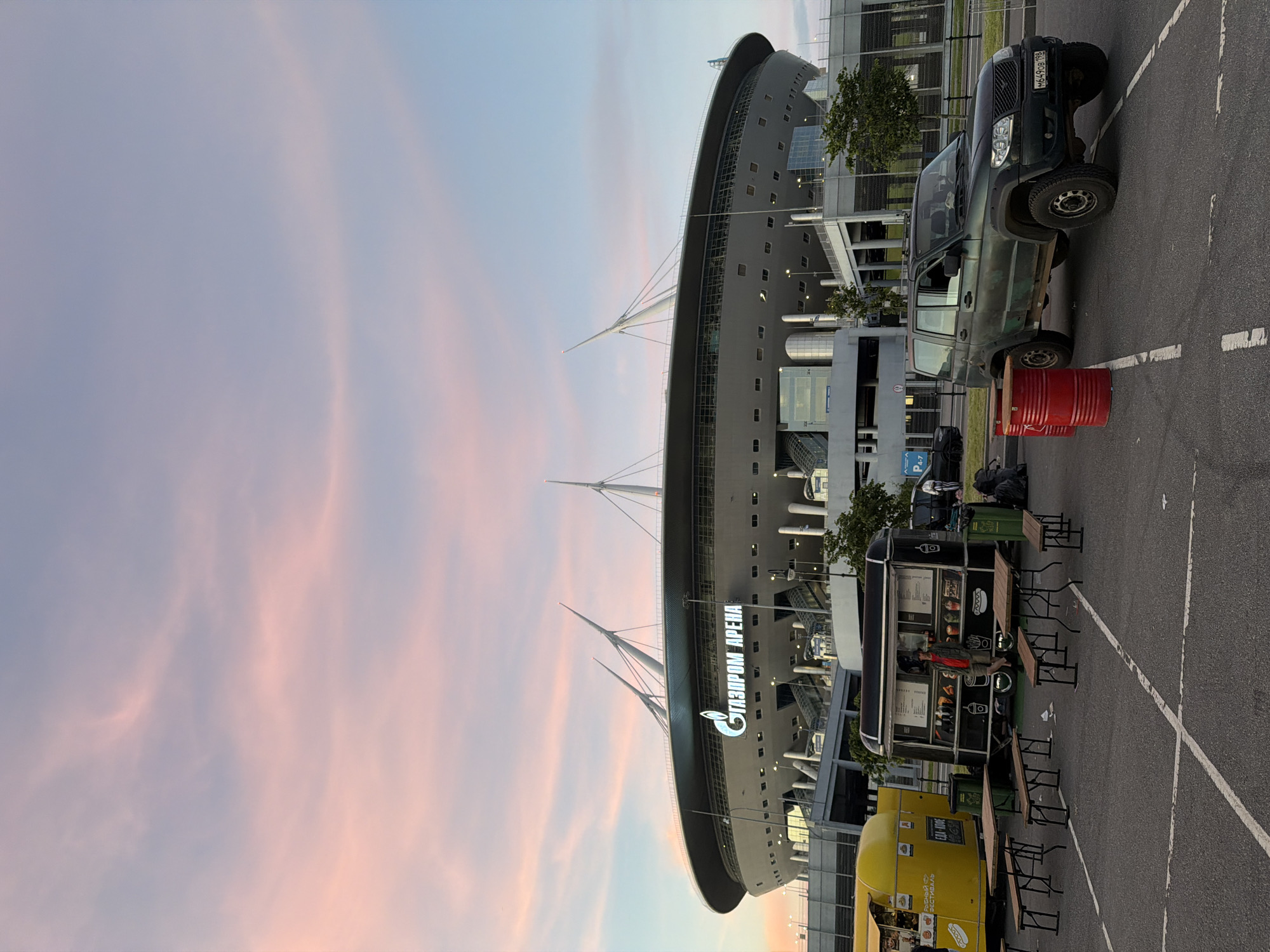 A wide shot captures a stadium in the background with "GAZPROM ARENA" in large illuminated letters. The sky above is a soft gradient of pink and blue, indicating either sunrise or sunset. In the foreground, a parking lot is partially visible, with two food trucks and several picnic tables. A dark green SUV is parked to the right, and two red barrels are positioned to the left of the vehicle. The scene appears to be outdoors, possibly during an event or gathering near the stadium.