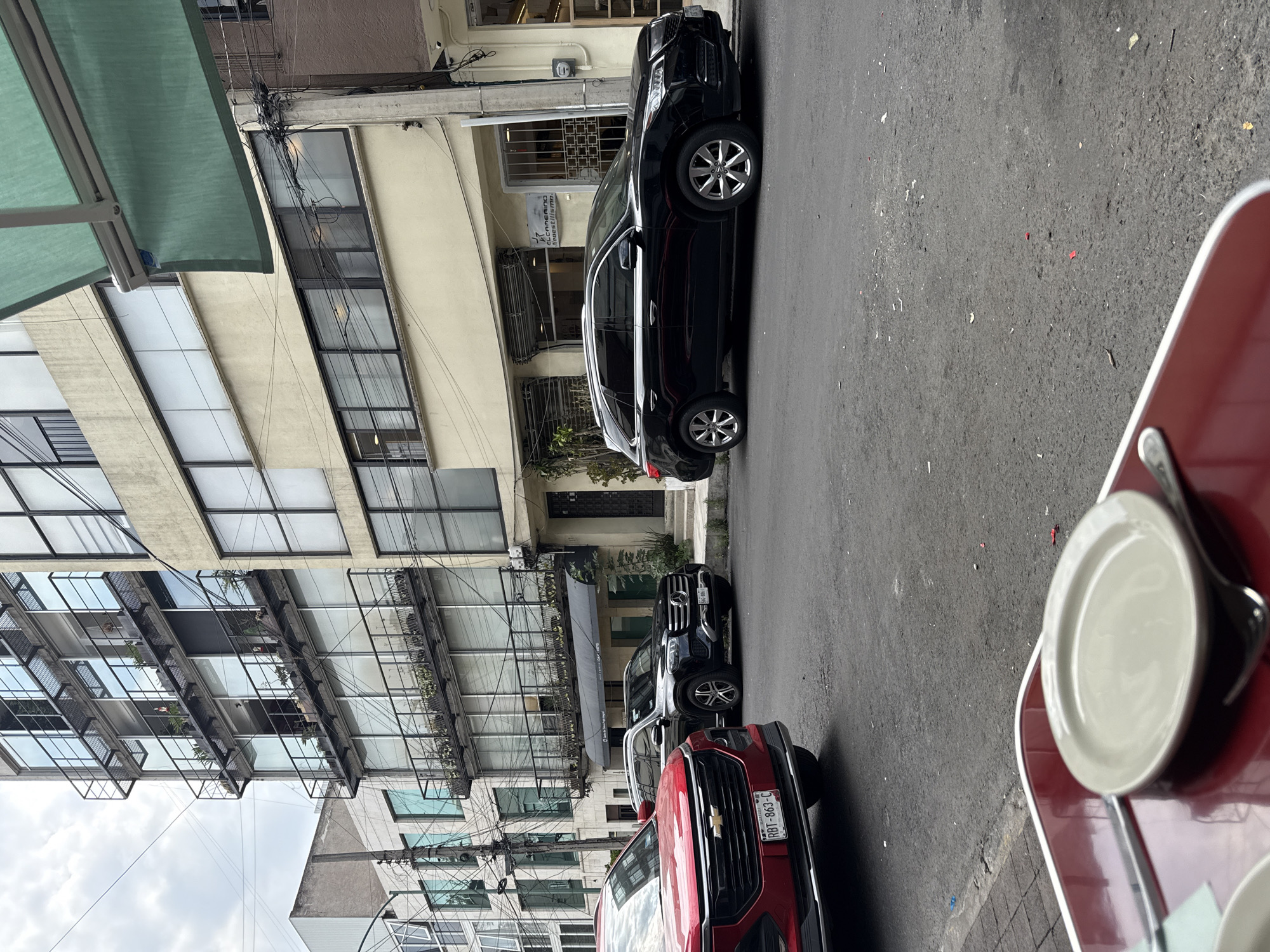 The image, taken from a slightly elevated perspective, likely from a cafe or restaurant patio, captures a daytime street scene in Mexico City under an overcast or partly cloudy sky. In the foreground on the right, a section of a red outdoor table is visible, featuring a beige plate and cutlery, indicating a dining setting. A green awning extends into the upper left corner, providing shade.
The street, a grey asphalt surface, stretches horizontally across the mid-ground. Several vehicles are parked along the curb. Closest to the viewer is a red Chevrolet Colorado pickup truck, displaying the license plate "RBT-863-C". Behind it is a black Mercedes-Benz GLC SUV with the license plate "906-YMJ". Further down the street, another black SUV, possibly an Acura, is parked.
On the left side of the street, multi-story buildings line the thoroughfare. One prominent building features a modern design with numerous large windows and glass balconies, some adorned with potted plants. Another building has a more classic facade, and a sign on it is partially visible, reading "EL CAMERINO". Numerous tangled power lines crisscross overhead, a characteristic feature of urban infrastructure. A reversed reflection of "BARBERIA" is also visible in a building's window. The overall scene depicts a typical, bustling urban environment.