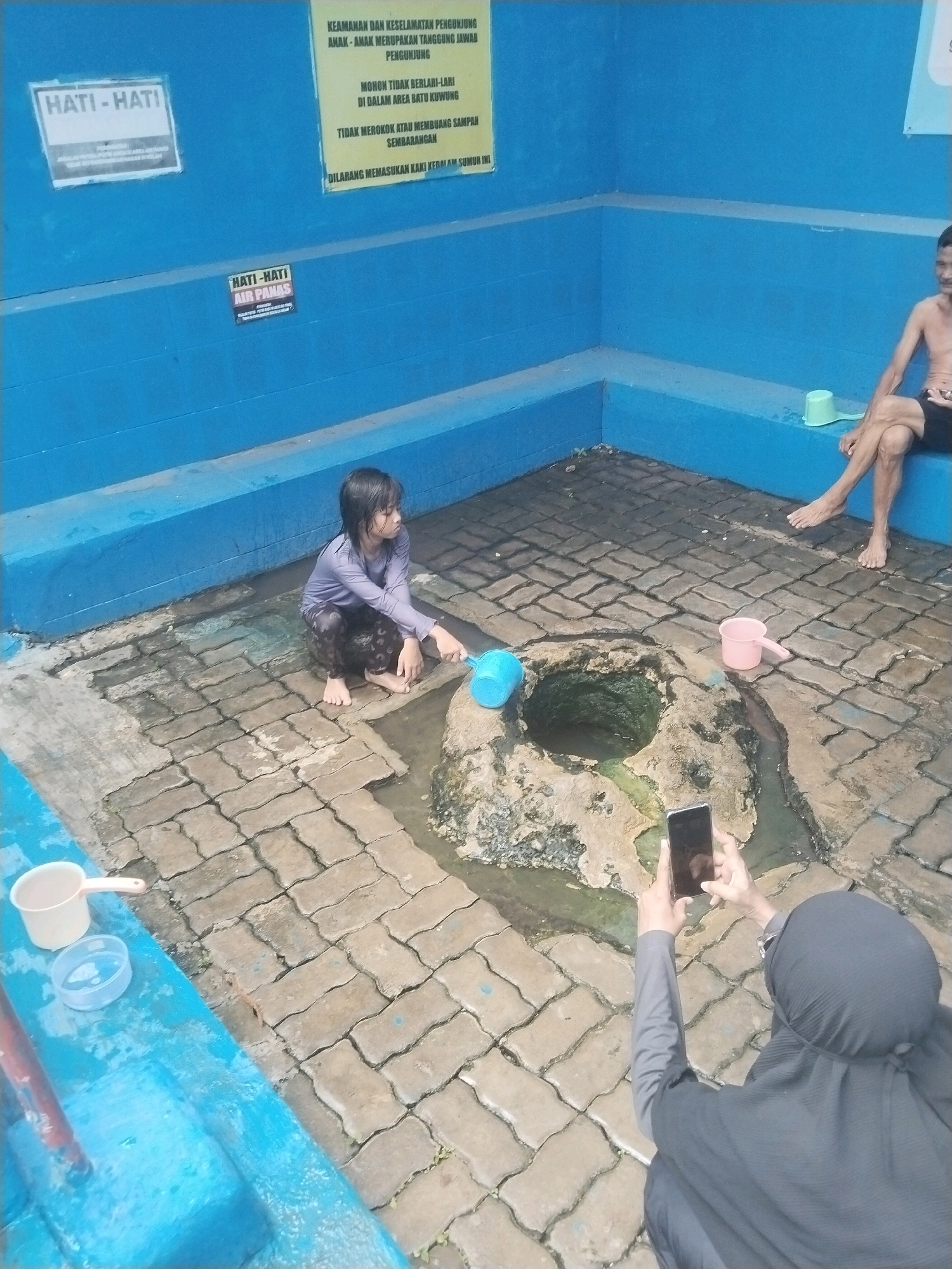 In the city of Batukuwung, Indonesia, a young girl with wet hair is crouched by a natural spring. She is wearing a purple long-sleeved shirt and dark patterned leggings, and she is holding a blue ladle in her right hand. The spring is surrounded by textured paving stones, and the area appears to be partially covered by water. To the right of the spring, a pink ladle rests near a rock. In the background, a man in black shorts is sitting on a blue ledge, with his legs crossed. Further to the right, a green ladle is visible on the ledge. On the blue wall behind the spring, there are two signs. One sign, partially obscured, says "HATI - HATI" (Be Careful). Another yellow sign contains Indonesian text, including "KEAMANAN DAN KESELAMATAN PENGUNJUNG" (Visitor Safety and Security). A person wearing a dark grey hijab and long-sleeved top is partially visible in the foreground, holding a smartphone and taking a photograph of the scene. White and clear plastic containers are also present on the blue ledge to the left.