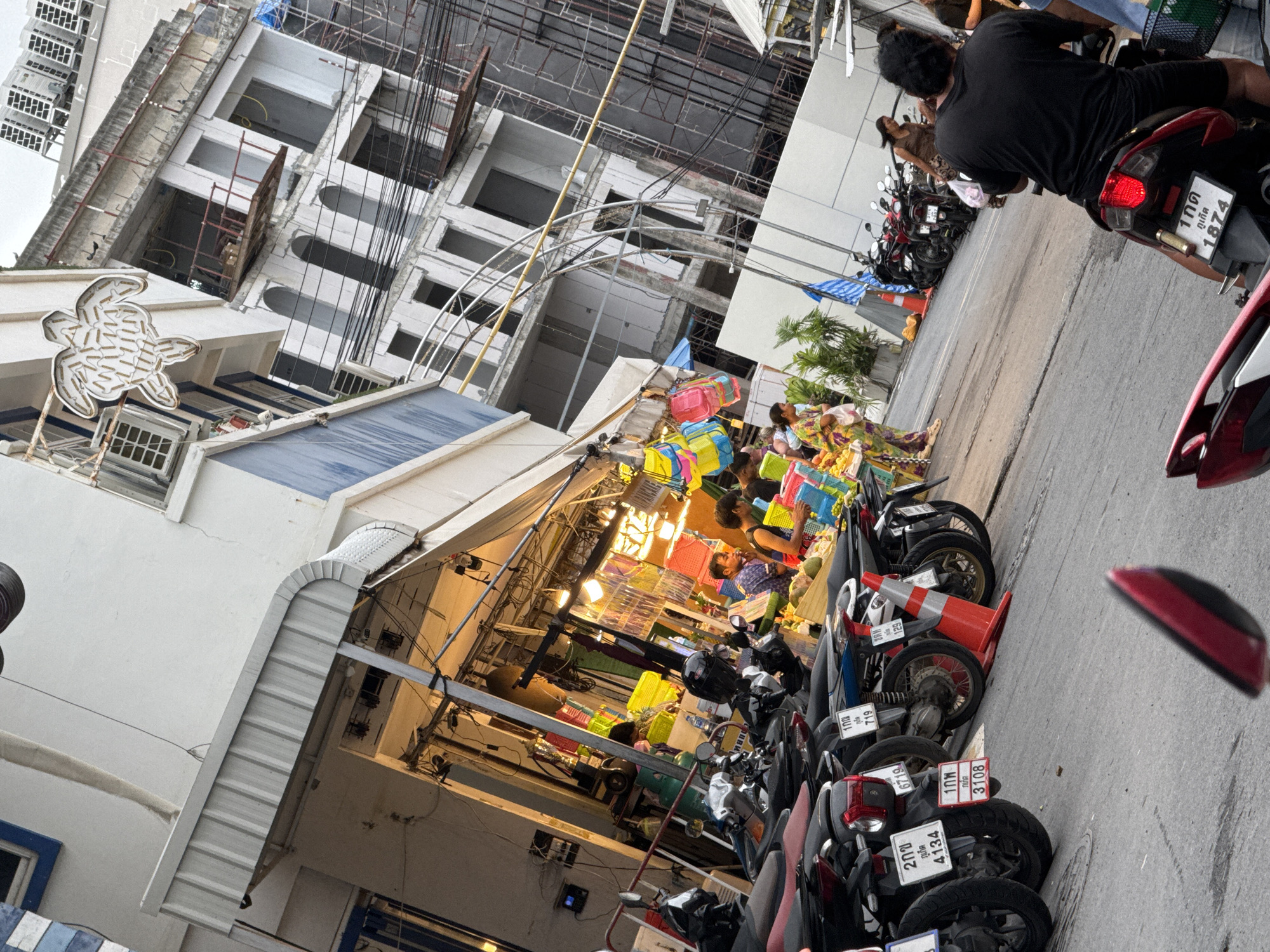 This media file captures a street scene in Karon, Thailand, likely during the daytime. The image is oriented such that the street runs diagonally from the bottom right to the top left. In the foreground and midground, a row of parked motorcycles is visible, some with Thai license plates. Behind the motorcycles, a market stall is set up, displaying an array of colorful baskets. People are present, some standing and interacting with the stall, while others are seated on or near the motorcycles.
The background features a multi-story building that appears to be under construction, with scaffolding and exposed structural elements. Overhead, a tangle of electrical wires crisscrosses the scene. The lighting suggests it could be late afternoon or early morning, with a slightly overcast sky. The overall atmosphere appears to be that of a busy local market or street vendor area. Visible text includes Thai characters on license plates, such as "ภูเก็ต" (Phuket) and various numerical designations.