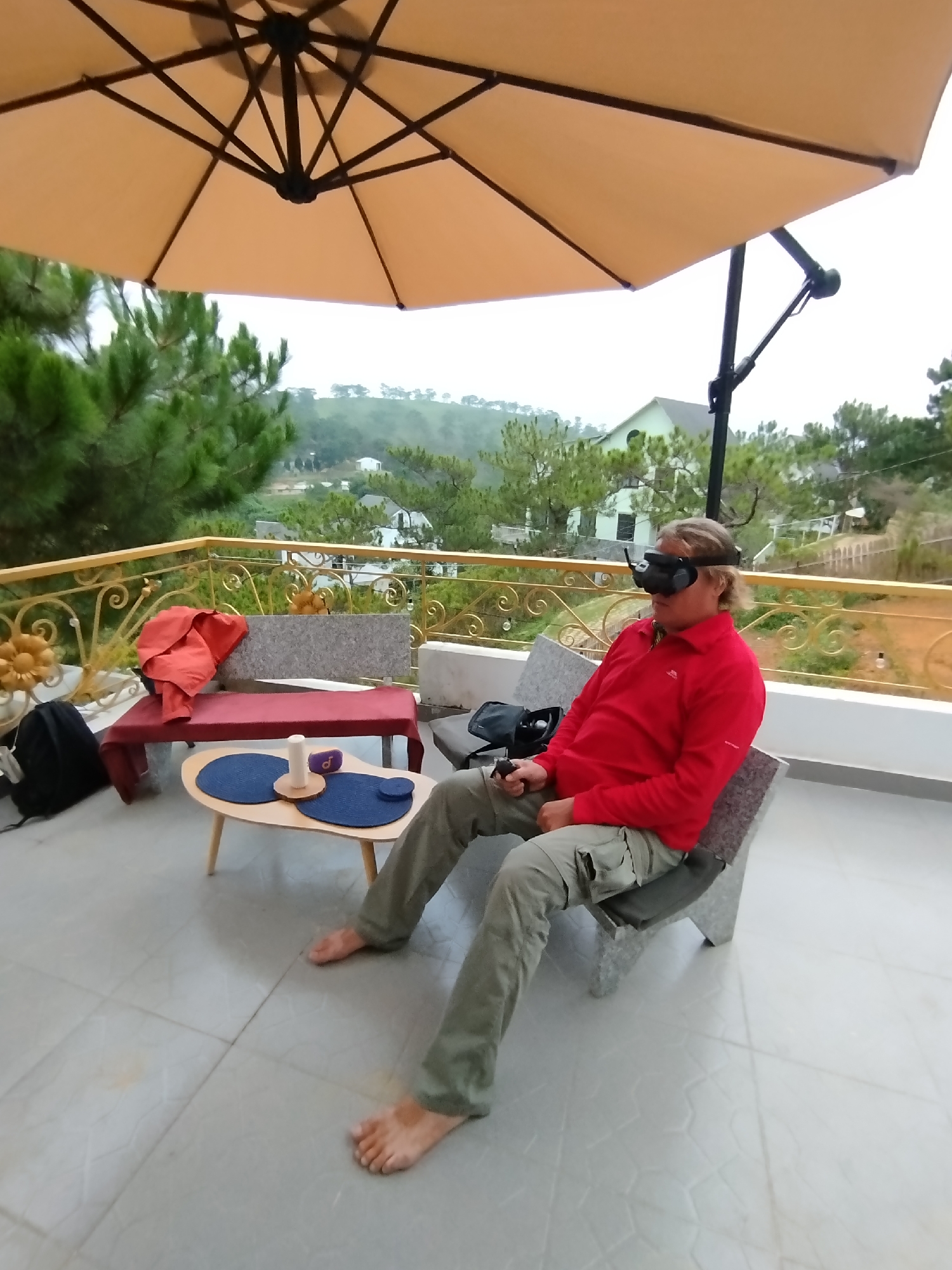 A man is sitting in a stone chair on a tiled patio, wearing VR goggles and holding a remote controller. He is barefoot and dressed in a red long-sleeved shirt and khaki pants. Above him is a large tan patio umbrella. Behind him is a gold-colored railing with green trees and a hillside with buildings visible in the distance. To his left is a small coffee table with a candle and other small objects on it, and a bench with a red jacket draped over it. A black backpack is on the ground near the bench. The location is Lạc Dương, Vietnam.