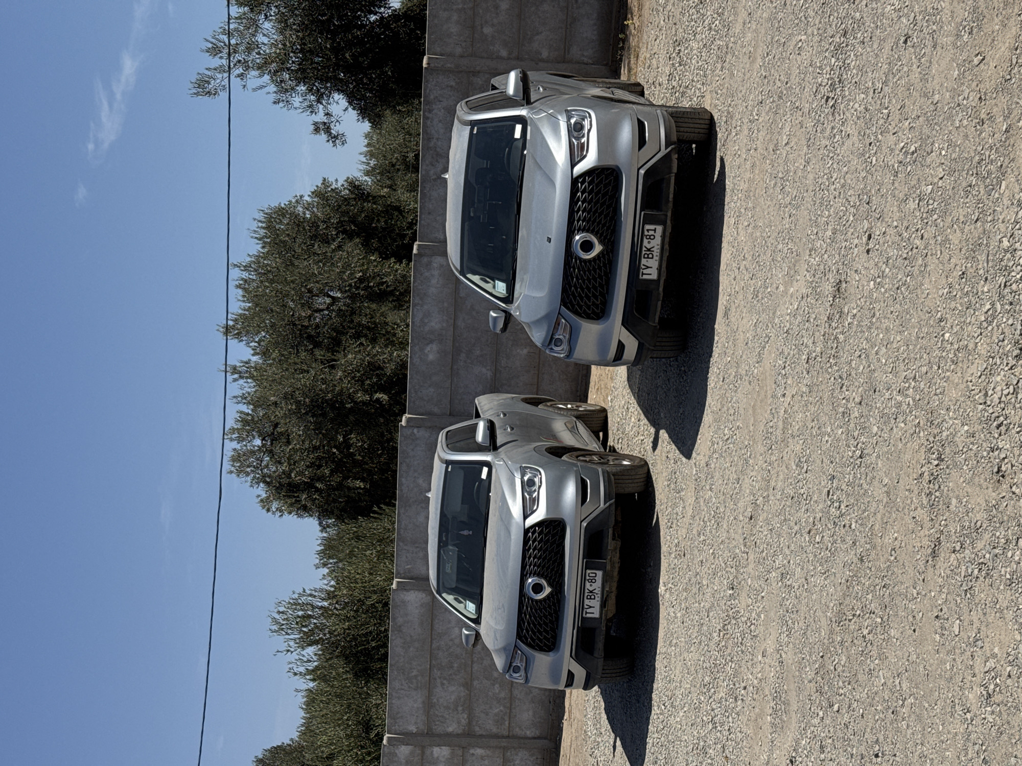 The image shows two silver SsangYong Actyon Sports pickup trucks parked on a gravel surface next to a concrete fence. Arica, Chile is determined by the license plates marked "TY BK-80" and "TY BK-81", as well as the country indication on the license plates. Trees are behind the fence, with a clear blue sky visible. No people are present.
