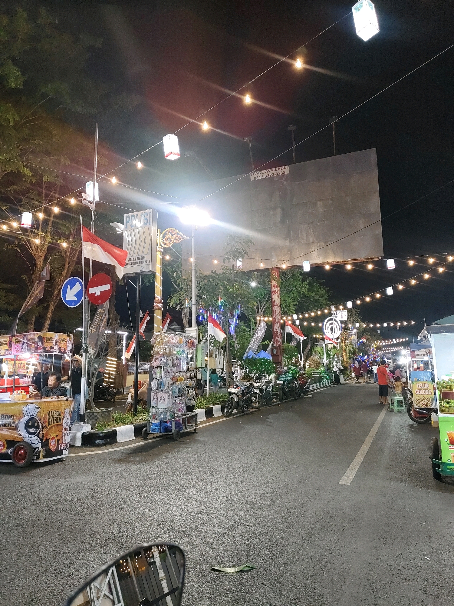 This is a nighttime street scene in Probolinggo, Indonesia, likely a bustling marketplace or event. The street is lined with stalls and food vendors, illuminated by strings of festive lights and decorative lanterns hanging overhead. Indonesian flags are prominently displayed, indicating a celebration or national holiday.
On the left side of the street, a food cart is visible with a cartoon character on its side and the text "MAKRONI NYEREM" and "GERO." A man is seated at the cart, possibly a vendor or customer. Next to this cart, a stall displays various items, including sunglasses and what appear to be religious trinkets, arranged on a rolling display.
Across the street, more vendors are set up, including one selling "TELUR GULUNG" (rolled eggs) and another with a display of green fruits, possibly avocados or coconuts. Several motorcycles are parked along the sidewalk, suggesting people arrived by bike.
In the background, there's a large, empty billboard with the text "SPACE AVAILABLE 0812-3322-8888." Signs in Indonesian are visible, including "POLISI" (Police) and "JALAN MASUK" (Entrance Road). People are walking along the street, browsing the stalls, and interacting with vendors. The overall atmosphere appears lively and festive.
