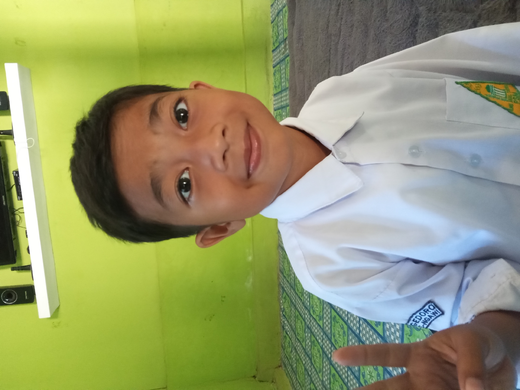 A young boy, appearing to be of Indonesian descent, is captured in a medium shot, posing with two fingers raised in a peace sign. He is wearing a white collared shirt, which has a patch on the left sleeve embroidered with the text "GEDORO NGANI" and another patch on the right chest with a green and yellow emblem. The boy has dark hair and dark eyes, and he is smiling directly at the camera. He is positioned in what appears to be a domestic interior, with a bright green wall behind him. In the background, a shelf is visible mounted on the wall, holding a black electronic device, possibly speakers or a television, and a white cable. The floor is covered with a patterned mat, featuring green and white geometric designs. To the right of the boy, a dark gray textured surface is visible, potentially a carpet or furniture. The overall setting suggests a casual, indoor environment, likely in the city of Kedunggudel, Indonesia, given the detail on the sleeve patch.