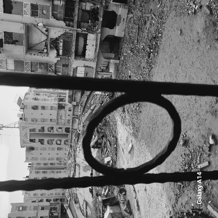A black and white shot through a metal railing captures a scene of urban devastation. Buildings appear heavily damaged, some with large sections missing. Rubble litters the ground. The photo suggests destruction from conflict or disaster, with the railing providing a stark foreground contrast. The view is slightly rotated, adding a sense of unease. "Galaxy A14" is visible in the bottom right corner.