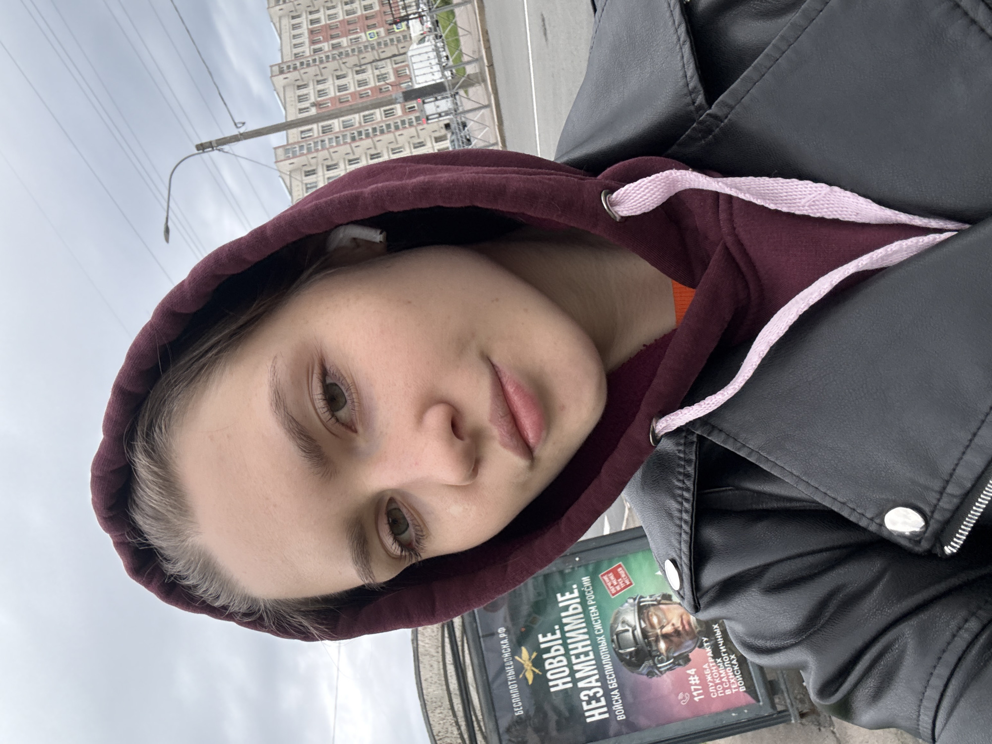 A young woman with light complexion and light brown hair, wearing a burgundy hooded sweatshirt with light pink drawstrings under a black leather jacket, poses for a selfie outdoors. She is looking into the camera.
Behind her, out of focus, is a street scene, including a multi-story building, possibly an apartment complex. A billboard features Russian text, an image of a soldier, and the URL "БЕСПИЛОТНЫЕВОЙСКА.РФ".
Based on the Cyrillic text and architectural style visible, the location is likely Saint Petersburg, Russia.