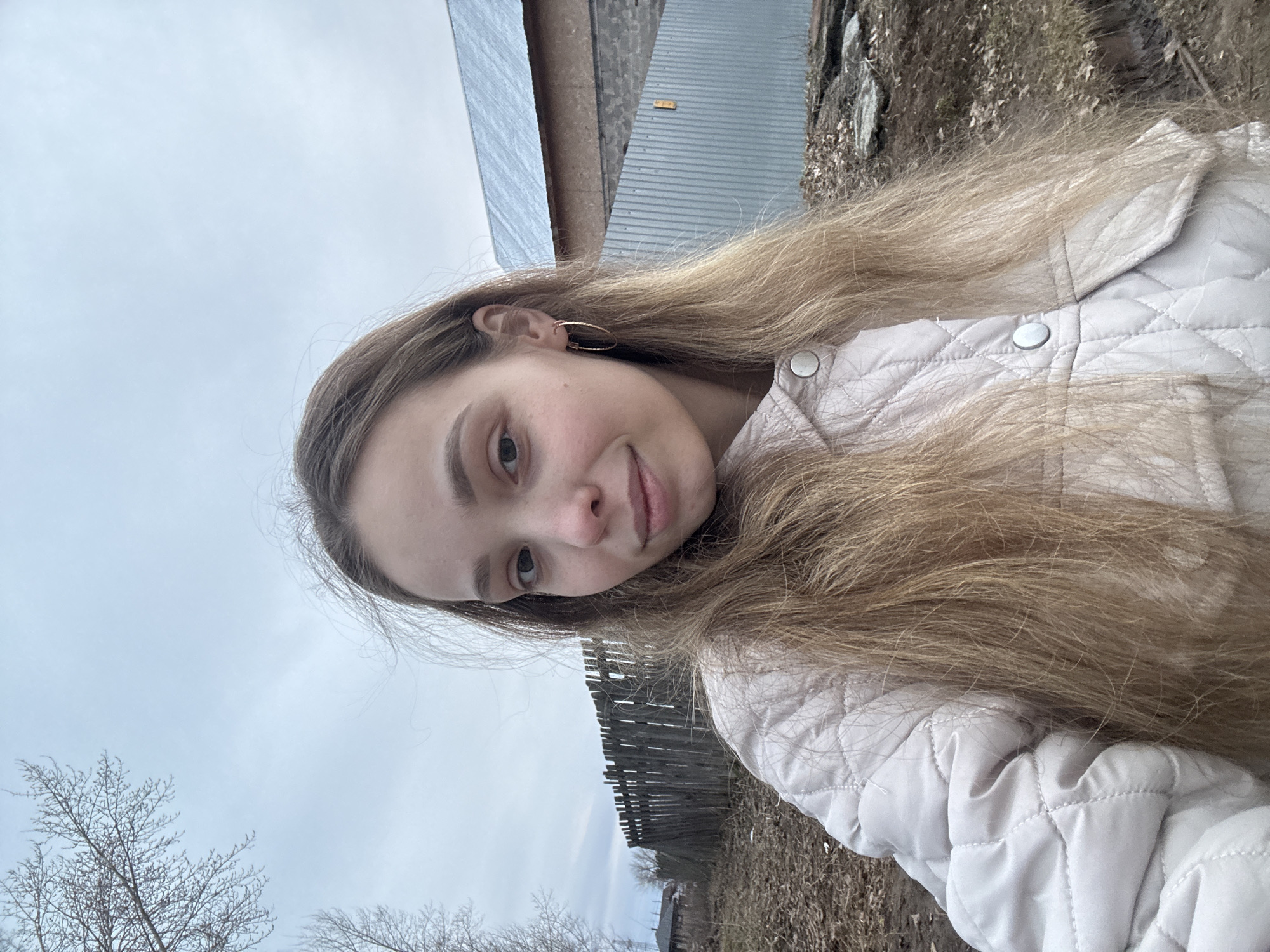A young woman is centered in the photograph, angled in a selfie-style shot. She has long, wavy brown hair and is wearing a light-colored quilted jacket with silver buttons and gold hoop earrings.
Behind her, there's a landscape featuring a gray sky and leafless trees, suggesting it could be early spring or late autumn. A wooden fence is partially visible. Buildings with corrugated metal roofing are in the background.
While the prompt mentions Chaikovsky, Russia, there is no way to verify this location from the image alone.
