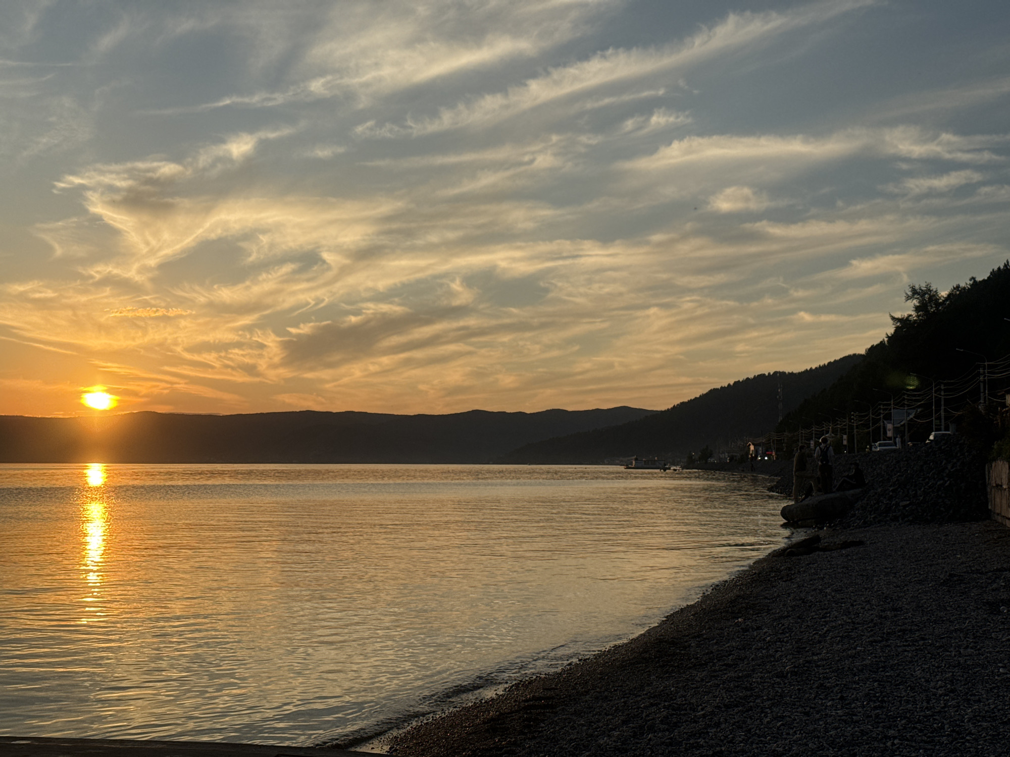 At sunset on the shore of Lake Baikal in Listvyanka, Russia, the sky is filled with wispy clouds, illuminated by the setting sun. The sun, a bright orb on the horizon, casts a golden reflection across the calm water. Silhouetted mountains form the background, and a tree-lined hill slopes down towards the right side of the image, where a dark, rocky shore meets the water. A few scattered pebbles lie on the shore, and a small boat is visible in the distance on the lake. Several figures are present on the shore near a cluster of rocks and trees, some appearing to be sitting or standing. Buildings and utility poles are visible along the hillside, suggesting a settled area.
