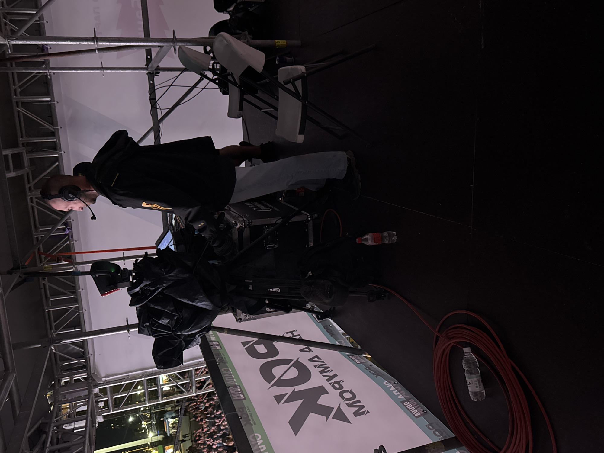 A man wearing headphones and a black jacket stands on a dark stage in front of a large white backdrop. He is positioned behind a large video camera and other broadcasting equipment. Two white folding chairs are visible to his right. In the background, a crowd of people can be seen, and in the distance, city lights and structures are illuminated. A large banner with the logo "BOX" is displayed prominently on the stage. A coiled red cable and a partially consumed bottle of water lie on the stage floor in the foreground. The setting appears to be an outdoor event or concert in the city of Khabarovsk, Russia, possibly at night.