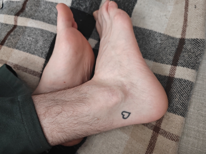 The image presents a close-up, high-angle view of two bare human feet resting on a plaid-patterned fabric, identified as a blanket or throw, exhibiting tones of beige, brown, and dark gray/black. The left foot and lower portion of the left leg are partially obscured by a dark green fabric, consistent with a trouser cuff, revealing visible hair on the exposed skin. The right foot, positioned slightly above and to the right of the left, displays a small, solid black outline heart tattoo on its outer ankle region. Both feet appear relaxed and stationary. The scene is located in London, United Kingdom.