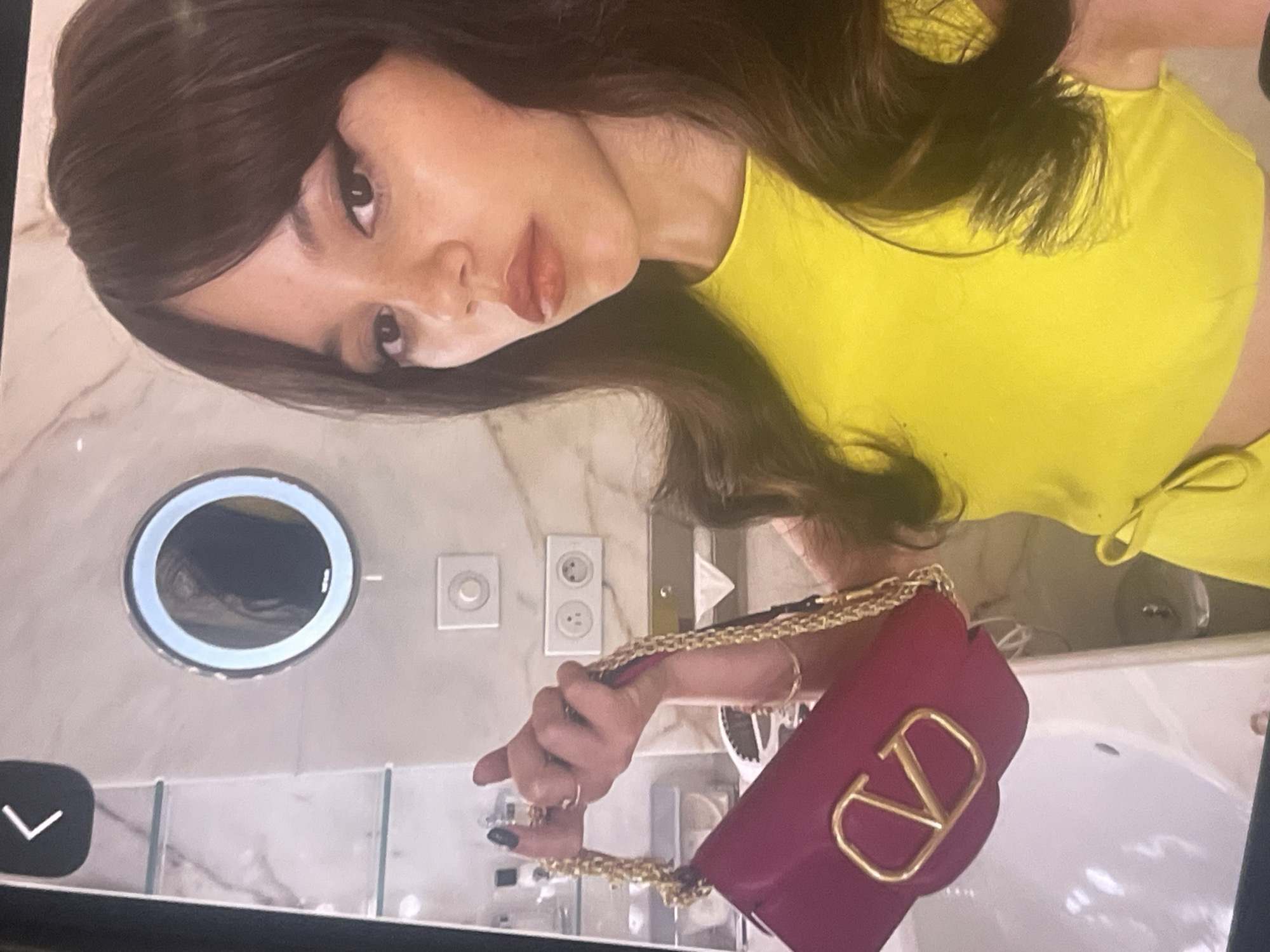 A young woman is depicted in a full-length, slightly tilted indoor portrait, likely a mirror selfie or a photo taken in front of a mirror, with the location context being Poltava, Ukraine. She has long, dark brown hair with bangs and is wearing a bright yellow, sleeveless top. Her facial features are accentuated with makeup, including dark eyeliner, well-defined eyebrows, and reddish-brown or nude lipstick, and she poses with a neutral, direct gaze.
She is holding a small, rectangular red handbag with a prominent gold "V" logo on its front, suggesting a Valentino brand. The bag features a gold chain strap, which she holds with her right hand, revealing dark-painted fingernails and rings on her fingers.
The background reveals a modern bathroom setting. A circular, illuminated mirror is mounted on the wall, partially reflecting the scene. Below it, there are a white light switch and two white electrical outlets on a wall covered with a light-colored, possibly marble-patterned, tile or wallpaper. A white sink is visible in the lower right, partially obscured by the woman and her handbag. The image is framed by what appears to be a device screen, featuring a black border and a white chevron arrow icon in the bottom-left corner. The lighting is consistent with an indoor environment, offering no specific cues regarding the time of day or weather.