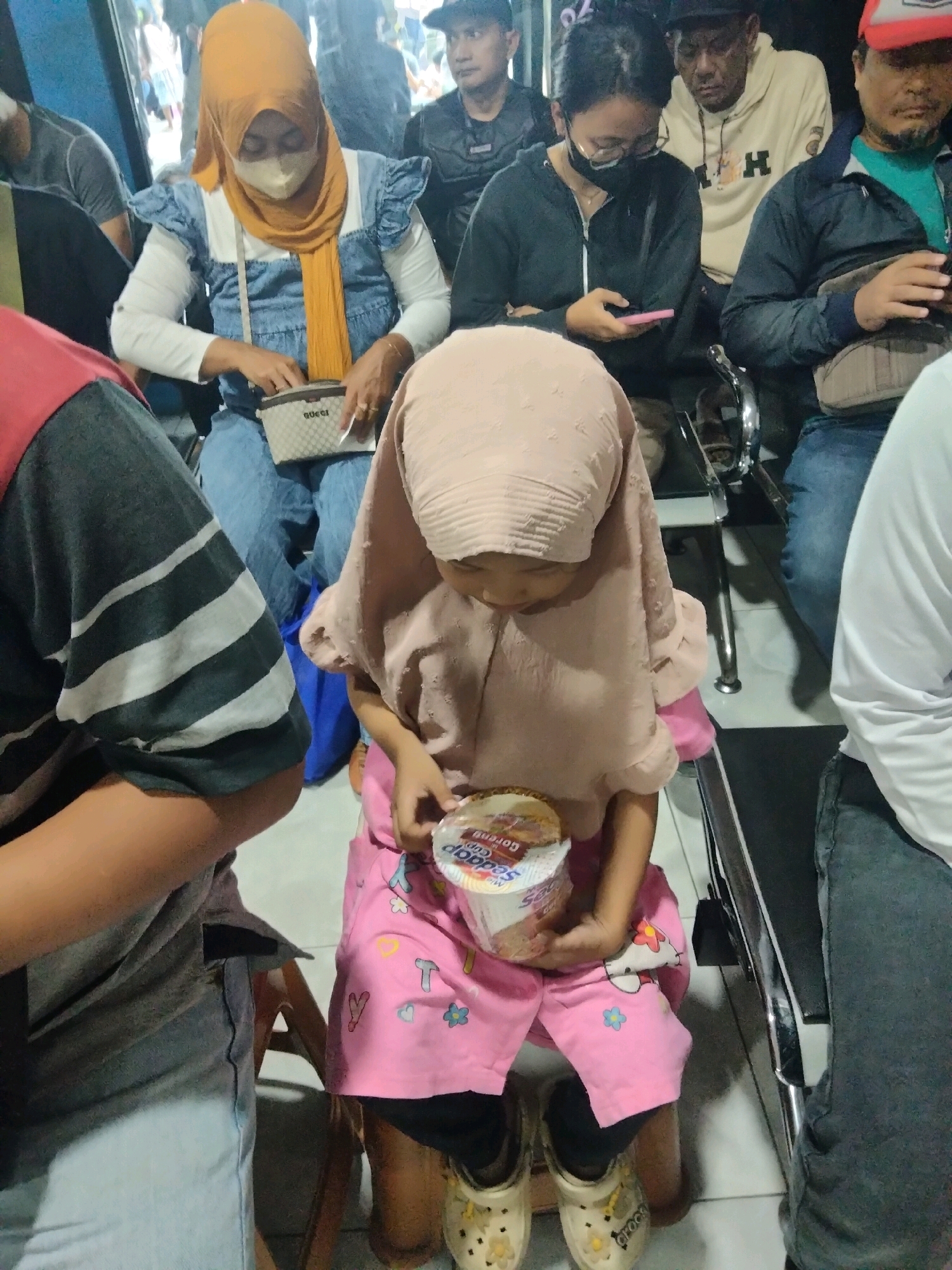 In the city of Sukaasih, Indonesia, a group of individuals are seated in what appears to be a waiting area, possibly a bus station or terminal. A young girl, wearing a pink hijab and pink patterned shorts, is in the foreground, holding a cup of instant noodles. To her left, a woman in a denim dress and orange hijab is rummaging through a small purse. Behind them, several other people are visible, some looking at their phones. The environment suggests an indoor setting with tiled flooring and seating areas.