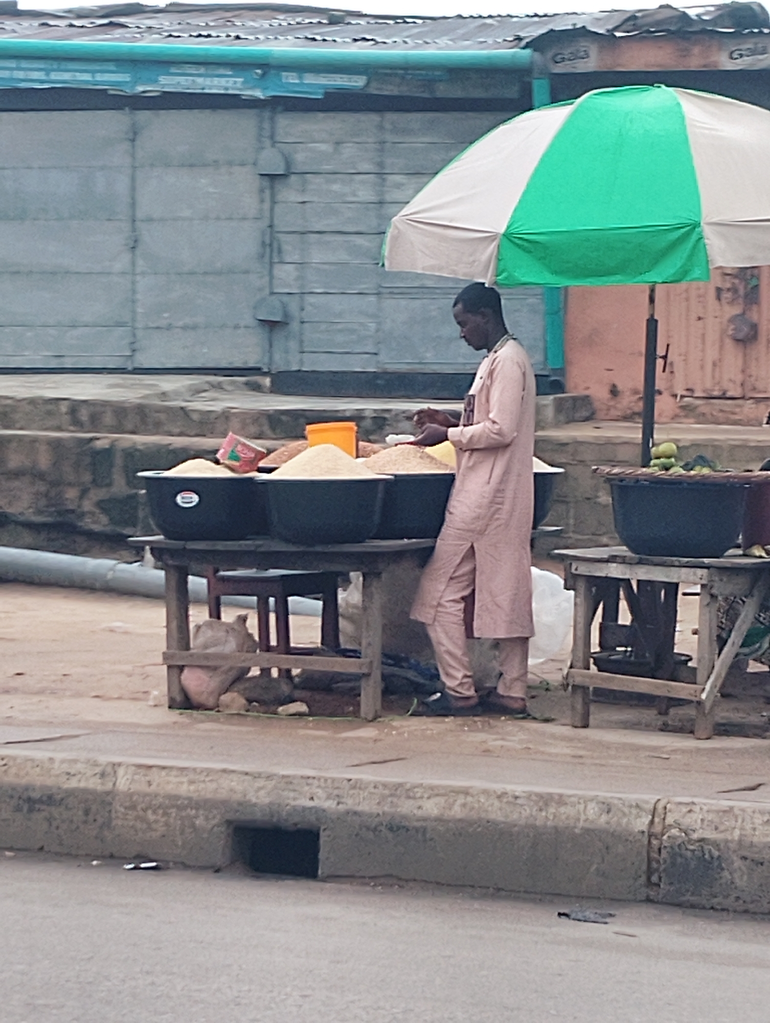 The image depicts a street vendor in Shangisha, Nigeria, standing under a green and white umbrella. The vendor, wearing a long, light-colored traditional outfit, is attending to a display of grains in large black basins. One basin is topped with a small, pink box. To the right, another table holds a woven basket filled with green fruit and a smaller, dark container. The vendor's stall is set up on a dirt ground, with a concrete curb and a road visible in the foreground. In the background, a building with large metal doors and a corrugated roof is present. The scene appears to be an outdoor marketplace or roadside stall.