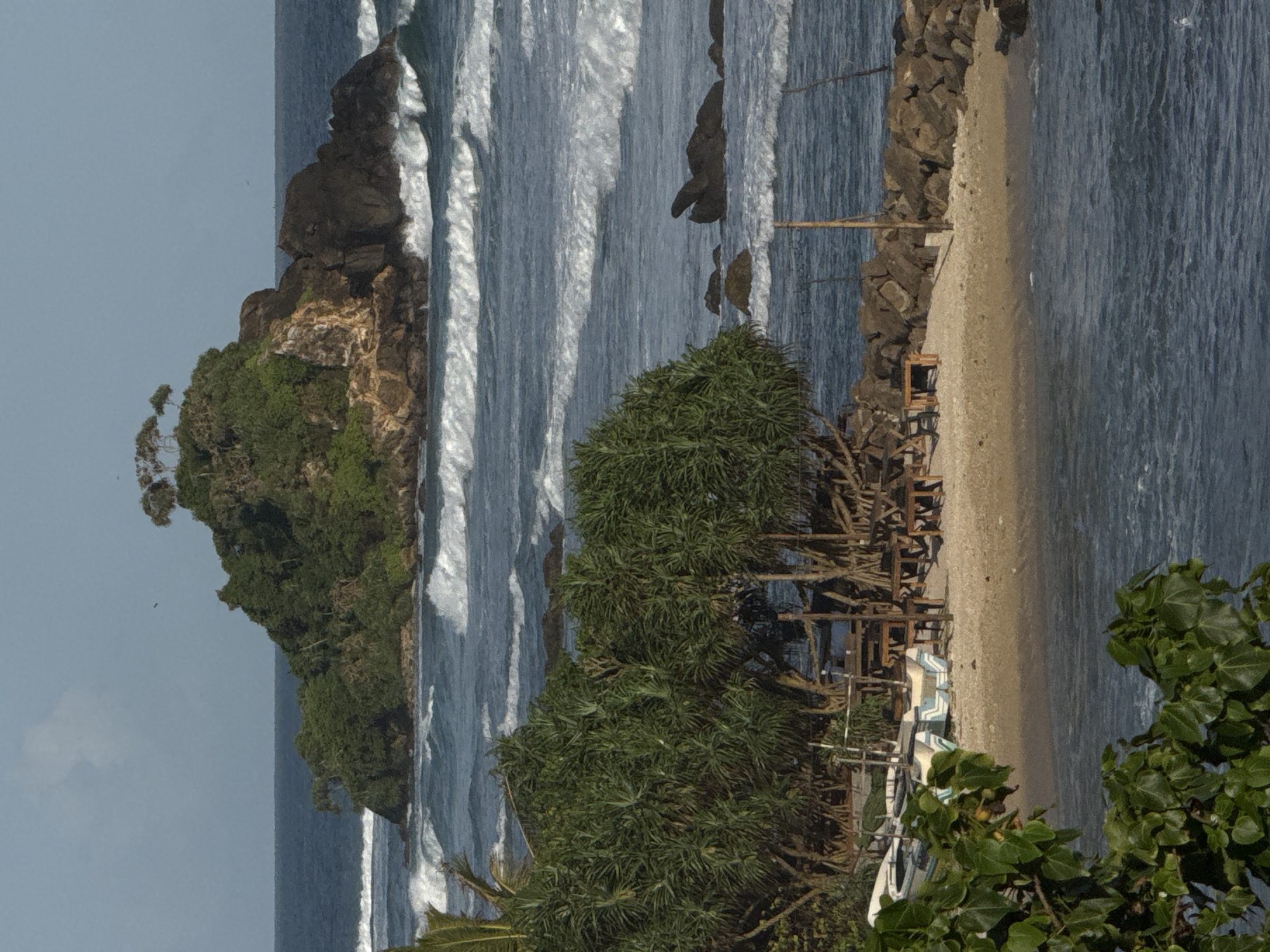 This is a photograph of a coastal scene in Goviyapana, Sri Lanka. The image captures a rocky island covered with lush green vegetation and a few trees in the distance, surrounded by the ocean with white-capped waves. In the foreground, there is a sandy beach with several wooden tables and chairs, suggesting a seating area for a restaurant or cafe. Two boats, one white with blue trim and another primarily white, are parked on the sand. A cluster of dense green trees with spiky leaves dominates the mid-ground of the image, with their roots exposed. The water is a deep blue, and the sky is a pale blue with a few wispy clouds. The overall impression is a tropical, serene beach setting.