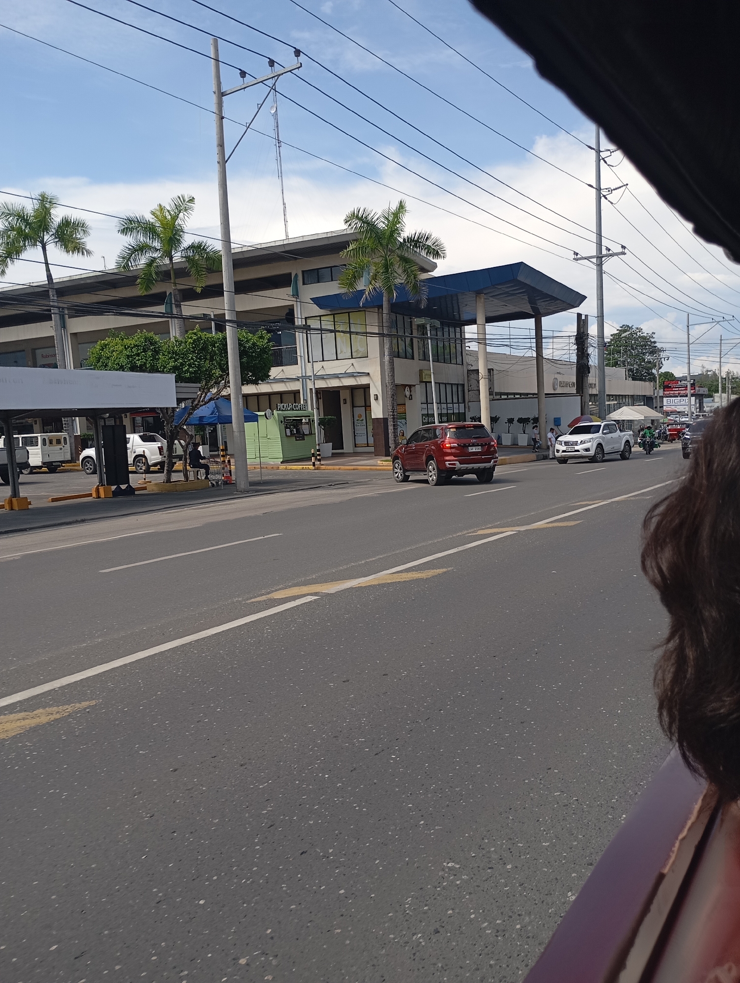 The image captures a bright, sunny daytime street scene in a commercial district of Davao City, Philippines, viewed from inside a moving vehicle, with a person's hair and shoulder visible in the foreground right.
At the center is a modern, multi-story commercial building, likely a shopping center or complex, characterized by a large blue-roofed entrance canopy and extensive glass facades. Partially visible text on the building facade suggests "Robinsons," while a smaller green kiosk in front reads "PICKUP COFFEE." Further down the street, another building bears the partial signage "UNION BANK." A person is seated on a bench under a blue tent near the "PICKUP COFFEE" kiosk.
The asphalt road, marked with white and yellow lines, is occupied by several vehicles. A red Ford Everest SUV with the license plate "LVF 2169" is prominent, followed by a white Nissan Navara pickup truck. Other cars, a white van, and tricycles are visible further down the road, indicating active traffic. Utility poles with a dense network of power lines extend across the sky. Palm trees and other green foliage line the sidewalks and the perimeter of the buildings.
The clear blue sky with scattered white clouds, combined with the strong sunlight, suggests it is likely late morning or early afternoon. Distant billboards, one showing "BIGPIX," are visible above the road. The overall scene depicts a typical, bustling urban environment in the Philippines.