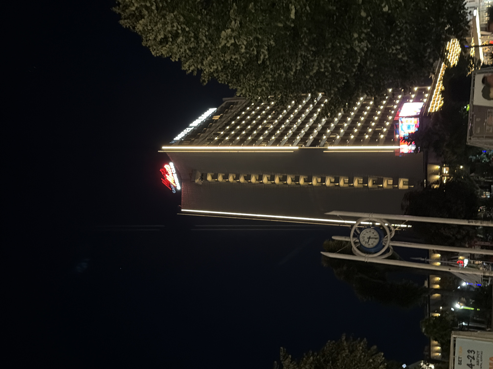 This is a nighttime, eye-level, medium shot of the Hotel Bulgaria in Burgas, Bulgaria. The hotel is a tall, modern building with numerous illuminated windows creating a grid pattern on its facade. A prominent red neon sign at the top of the hotel reads "GRAND CASINO CLUB". To the left of the hotel, another building is partially visible, also illuminated. In the foreground, a white clock tower with a blue clock face stands in the lower-left quadrant of the image. Trees with dark green foliage are visible on the right and left sides of the frame, framing the hotel. The sky is a deep, dark blue, indicating nighttime. The scene appears to be an urban setting at night, possibly a public square or street in front of the hotel.