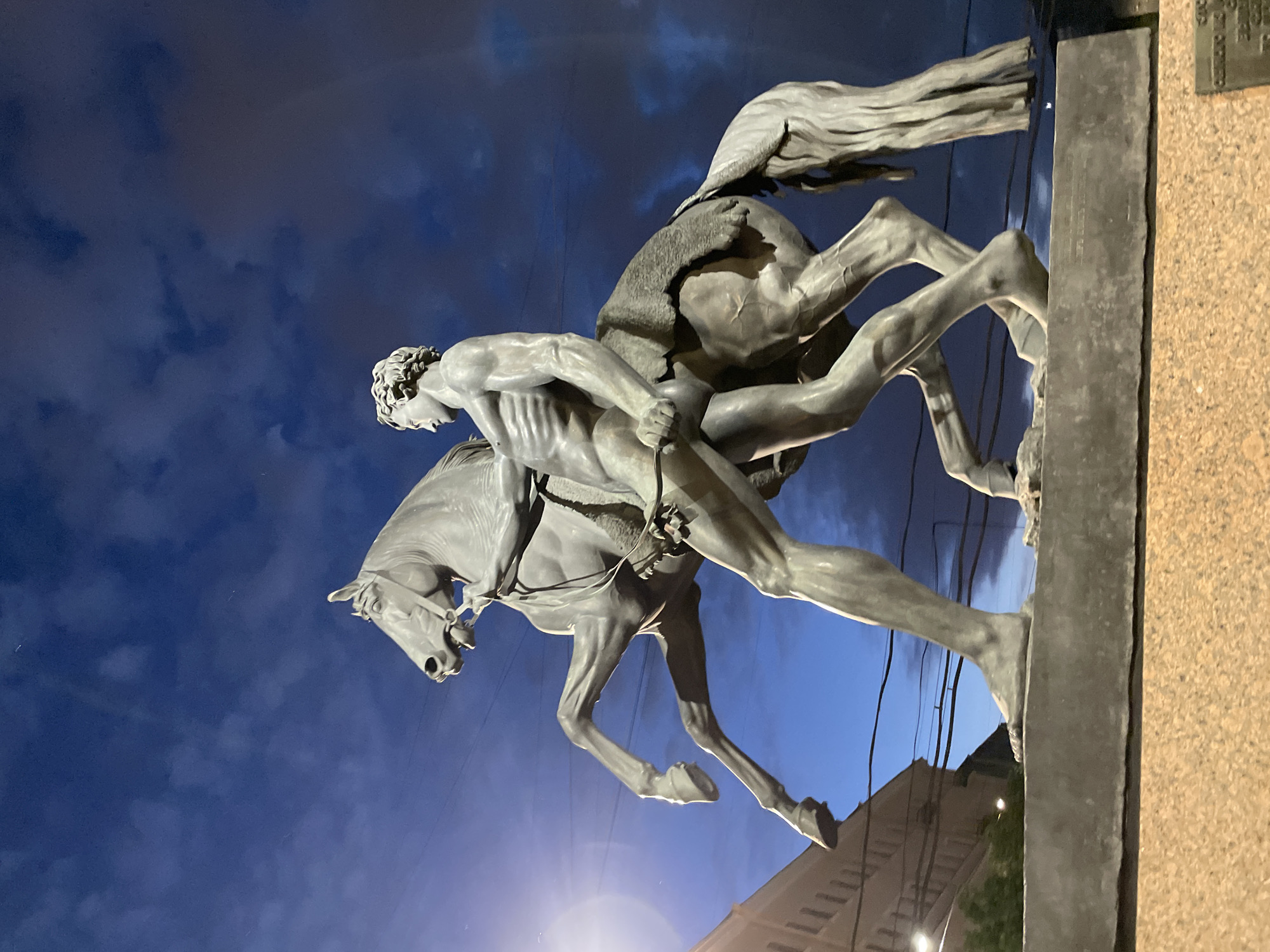 This is a low-angle, eye-level shot of the equestrian statue of Peter the Great, also known as the Bronze Horseman, located on Senate Square in Saint Petersburg, Russia. The statue is made of bronze and depicts Peter the Great on a rearing horse, trampling a serpent. The sky behind the statue is a deep blue with scattered clouds, illuminated by artificial lights from the city. The base of the statue is made of granite. In the background, buildings and power lines are visible, suggesting an urban environment. The time of day appears to be dusk or night due to the dark sky and artificial lighting.