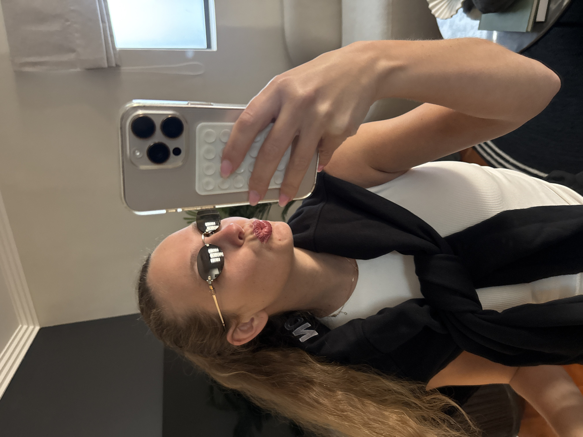 The image is a selfie taken by a young woman, likely reflected in a mirror, showing her from the chest up. She has light brown, wavy hair and is wearing stylish sunglasses with round, dark lenses and thin gold frames. Her lips are slightly pursed in a playful pout, covered in a glossy red or berry-toned lipstick.
She is dressed in a white ribbed tank top and has a black garment, possibly a sweatshirt or jacket, tied around her shoulders or neck, featuring a partially obscured white logo or text. Her left hand is visible, holding a light-colored smartphone with a clear case and a distinct white, rectangular PopSocket-like accessory on its back, featuring a grid of small circular studs. The phone's triple-lens camera system is prominent. Her fingernails are painted a light pink.
The background suggests an indoor setting, likely a room with light-colored walls. A window reflection is visible in the upper part of the image, indicating natural daylight. Indistinct objects like furniture (possibly a couch) and decorative items are blurred in the background. Part of another black garment with white stripes, possibly athletic wear, is visible on her lower body or in the background. The overall mood is casual and confident. While the image itself doesn't provide specific geographical cues, it is set in Cape Town, South Africa according to the provided context.
