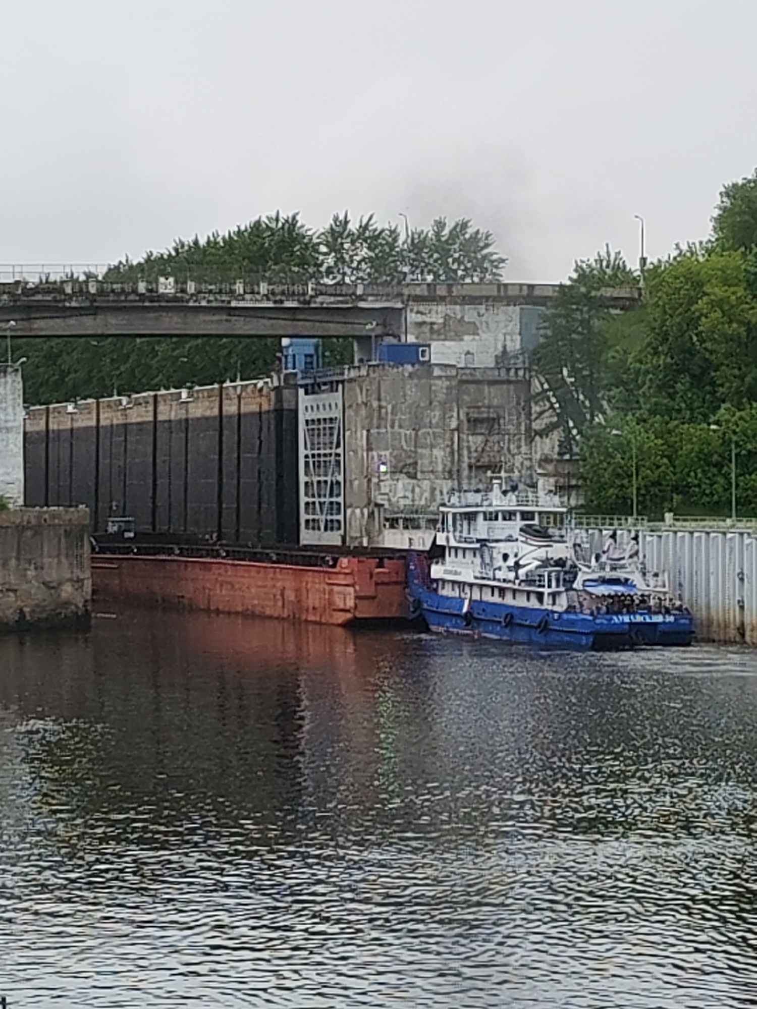 The image depicts a waterway scene in Bol'shie Mkhi, Russia. A blue and white passenger boat labeled "АМУРСК-30" is docked alongside a red barge within a concrete lock system. A pedestrian bridge spans the waterway overhead, lined with greenery. Additional trees and buildings are visible on both sides of the canal. The sky appears overcast.