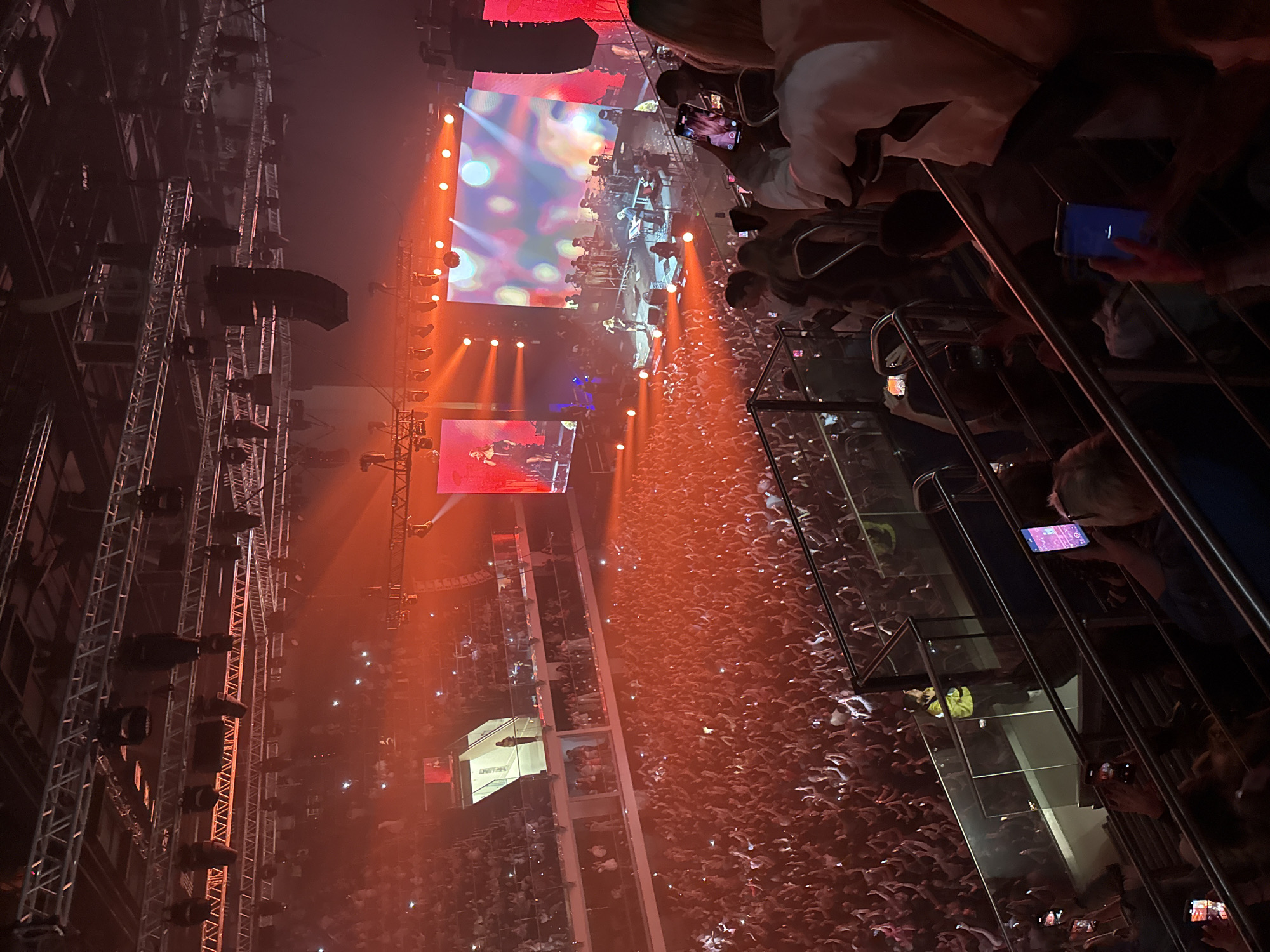 The image depicts a concert venue filled with a large crowd of people. The stage is visible in the distance with screens and lighting systems. Many attendees are holding up their phones, presumably recording or taking pictures of the performance. The venue is illuminated by orange and red lights. People are standing close together, indicating a densely packed audience. The viewpoint is from an elevated position, possibly a balcony or upper level.
