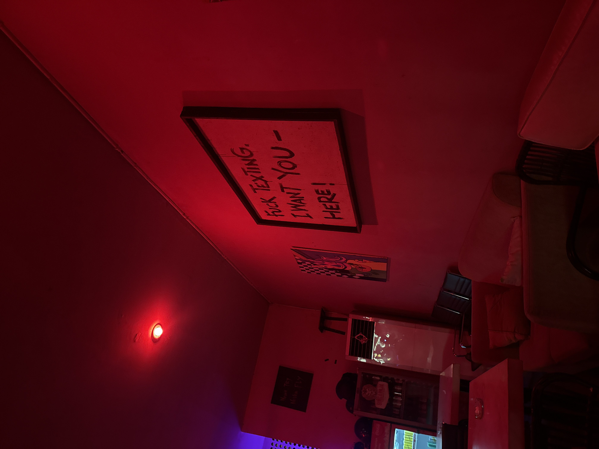 The image shows the interior of a bar located in North Kuta, Indonesia. The room is bathed in red light. On the wall is a framed sign that reads "FUCK TEXTING. I WANT YOU - HERE!" There are other framed images on the wall as well. A white couch is partially visible in the upper-right corner of the image. Below the signs on the wall is a refrigerator, partially visible. Some tables and chairs are also in view.