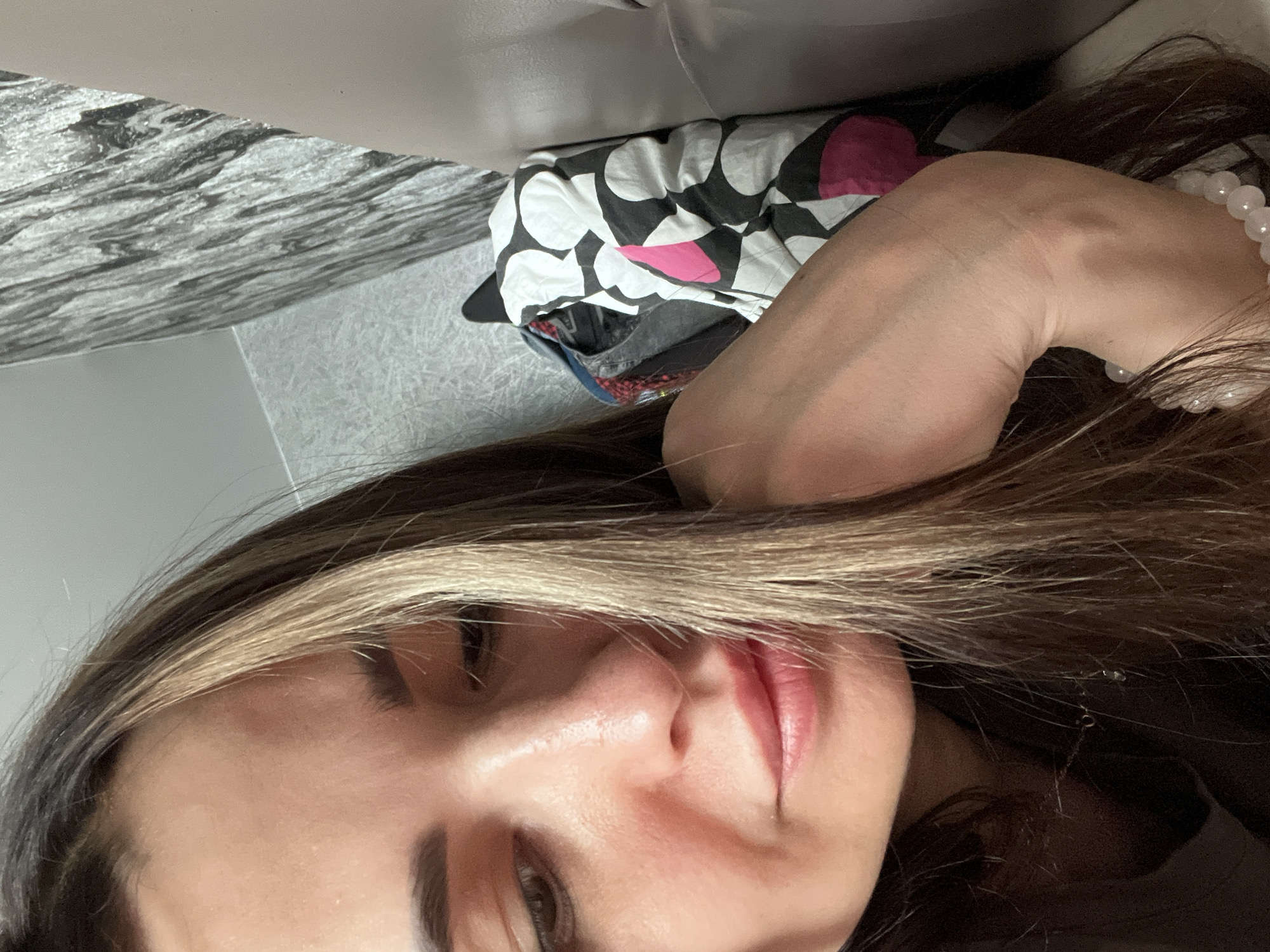 A close-up, rotated selfie shows a person, likely female, in a relaxed, reclined position indoors. The image is oriented with the subject appearing to be on her right side, looking upwards from the bottom left of the frame. Her face is partially obscured by long, dark brown hair with prominent blonde highlights.
Notable facial features include a visible dark eye, well-groomed dark eyebrows, a nose, and full lips with pink-red lipstick, showing a gentle, subtle smile. Her skin appears clear and smooth.
Her bare right arm and shoulder are visible on the right side of the frame, bent at the elbow, suggesting she might be resting her head or propping herself up. A white beaded bracelet, possibly made of stone or pearl, is worn on her wrist.
In the background, behind her head and arm, a crumpled pillow or cushion with a distinctive black, white, and pink geometric pattern (resembling circles or hearts) is visible. To the left, there's a wall or large surface featuring a black and white marbled or wavy textured design. Below that, a lighter grey or white textured surface, possibly fabric or carpeting, is present. The overall setting suggests an indoor environment, likely a bedroom or living room, with a relaxed atmosphere. There is no visible text in the image.