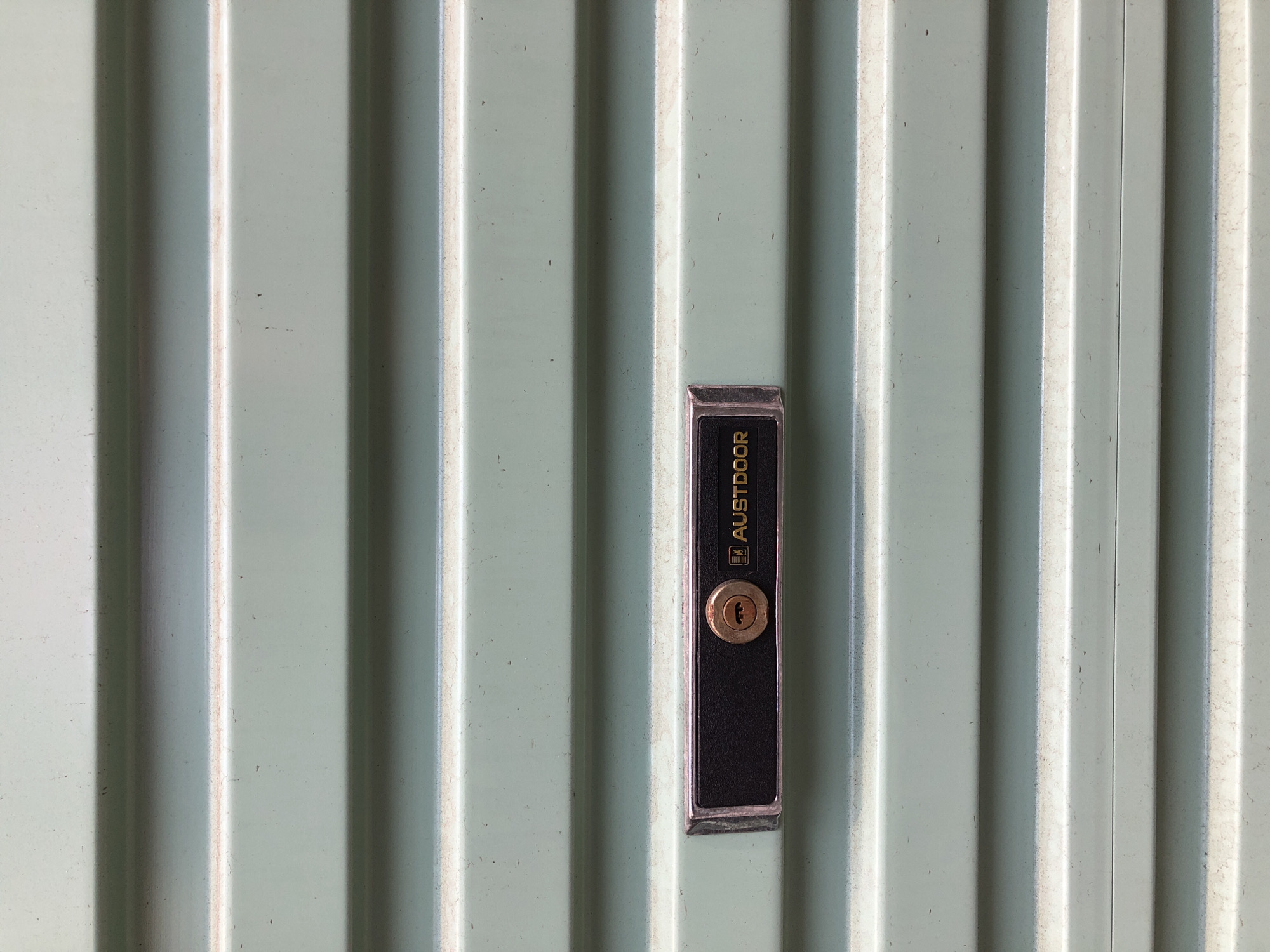 The image shows a close-up, head-on view of a ribbed, pale green metal surface, possibly a garage door or a similar industrial or security door. The surface is characterized by vertical indentations and raised ridges that create alternating areas of light and shadow. In the center of the frame, a rectangular black lock mechanism is mounted. The lock has a metallic silver trim around its edges and a circular brass keyhole at the bottom. Above the keyhole, the word "AUSTDOOR" is printed in gold lettering, with a small logo to the left of the text. The overall impression is one of a functional, outdoor element, likely in a residential or commercial setting. The lighting suggests it's daytime, but the specific time or weather conditions are not discernible. There are no people or other objects visible in the frame. The texture and material of the door suggest it is exposed to the elements. Given the location context of Da Nang, Vietnam, this could be a common feature on many types of doors in the region.