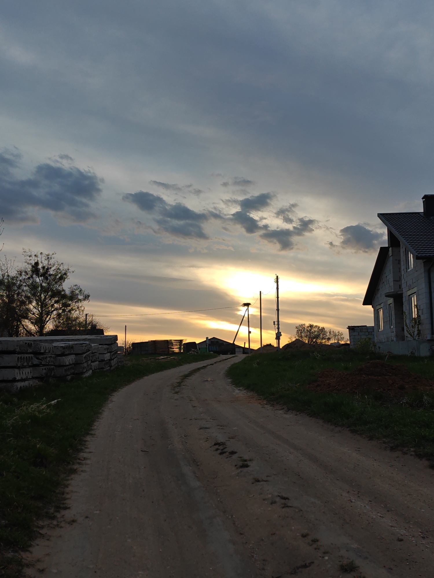 The image captures a rural or semi-rural landscape at sunset or dusk. A winding, unpaved dirt road dominates the lower center, curving gently to the left and receding into the distance. To the left of the road, a large stack of grey concrete slabs, likely building materials, sits on a grassy verge. Behind the slabs, a tree with sparse foliage and visible mistletoe-like growths stands against the sky.
On the right side of the road, a grassy bank rises, featuring a mound of dark soil. A partially constructed house with light-colored block walls and a dark grey tiled roof is visible, with windows catching some of the lingering light. In the middle distance, towards the setting sun, several utility poles are silhouetted. One pole prominently features a large bird's nest at its top, likely belonging to a stork.
The sky is a significant element, displaying a mix of dark grey clouds overhead transitioning to warmer yellow, orange, and light grey hues at the horizon where the sun is setting, casting a radiant glow. The overall impression is one of a quiet, transitional moment at the end of the day, with signs of ongoing development in a natural setting. There are no people or discernible text in the image. The scene suggests a location like Путрышкаўскі сельскі Савет, Belarus, fitting with the rural character and presence of a potential stork's nest.
