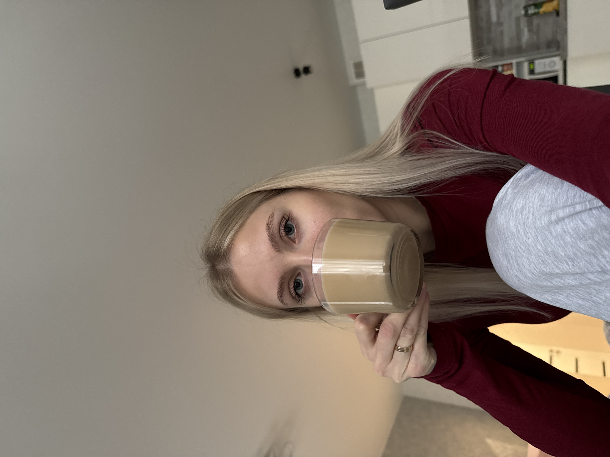 A woman with blonde hair is holding a glass of coffee or a similar beverage in front of her face. She is wearing a burgundy long-sleeved shirt and gray sweatpants. The background includes a light-colored wall and a glimpse of kitchen cabinets. The image appears to be taken indoors, and there is no indication of being in Izhevsk, Russia.