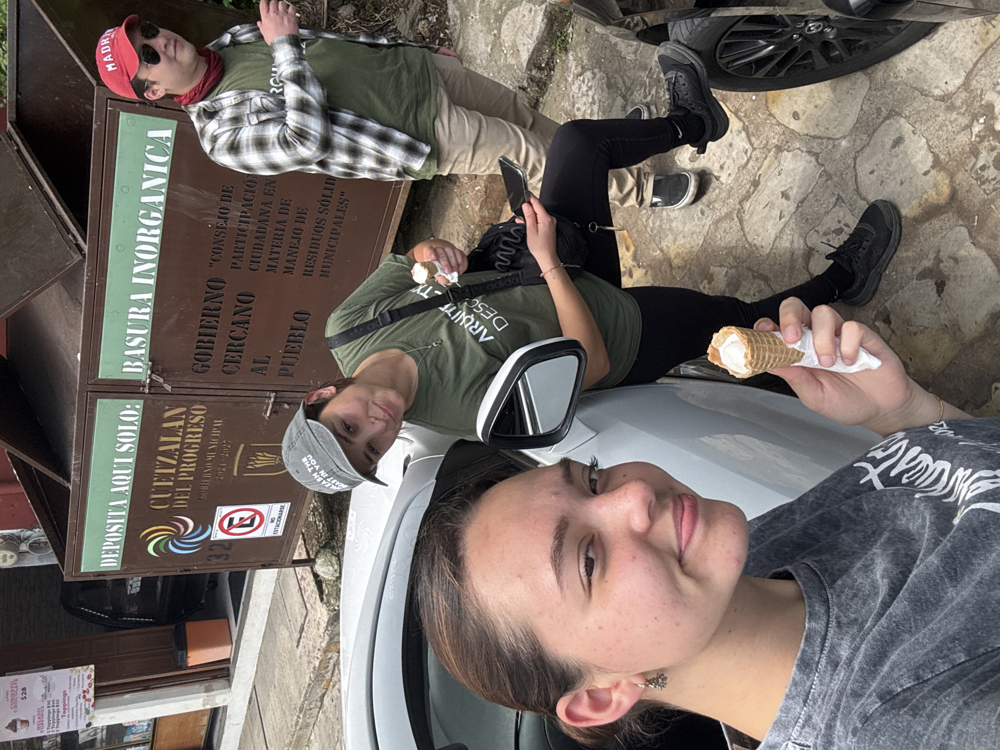The image appears to be a selfie taken by a person with short brown hair, wearing a gray t-shirt. She's standing in front of a white car and holding an ice cream cone. Beside her, another person in a gray hat and green shirt is leaning against the car, holding a phone. In the background is a third person, wearing a red hat, sunglasses, and a checkered shirt, reclining on top of a brown metal trash container. The trash container is labeled in Spanish, including "BASURA INORGANICA" and "CUETZALAN DEL PROGRESO", indicating the location in the city of Cuetzalan, Mexico. The trash container also has signs for the "Consejo de Participacion Ciudadana en Materia de Manejo de Residuos Solid Municipales". A "No Parking" sign is also visible on the trash bin. A sign advertises "Cripto Crepes" with "Helado Toppings" and various toppings for an added charge. The ground is paved with stone.