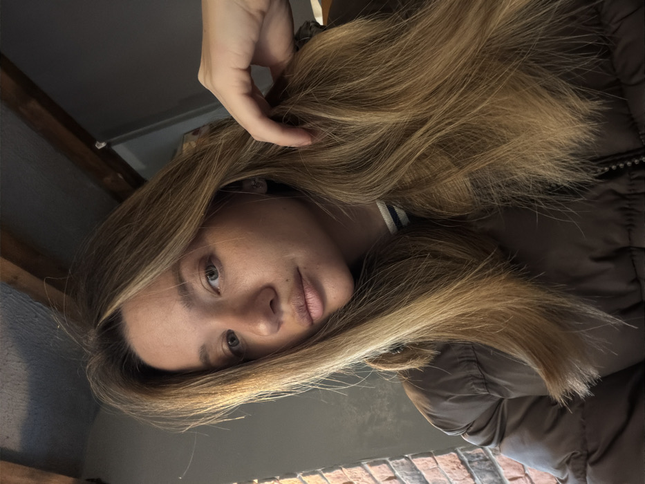 A close-up, slightly low-angle photograph depicts a female individual indoors in an unspecified location in Unknown, Unknown. The subject, with light skin and long, multi-tonal blonde hair, looks directly forward with a neutral expression. Her right hand is visible on the right side of the frame, fingers loosely gripping or resting in her hair. She wears a brown puffer jacket, partially zipped, over a dark garment with a white collar featuring two horizontal dark stripes. A small, light-colored stud earring is visible in her right ear. The background on the left consists of a reddish-brown brick wall. The upper background features a dark ceiling or wall with exposed wooden beams, and a dark, featureless wall or partition is visible on the right.