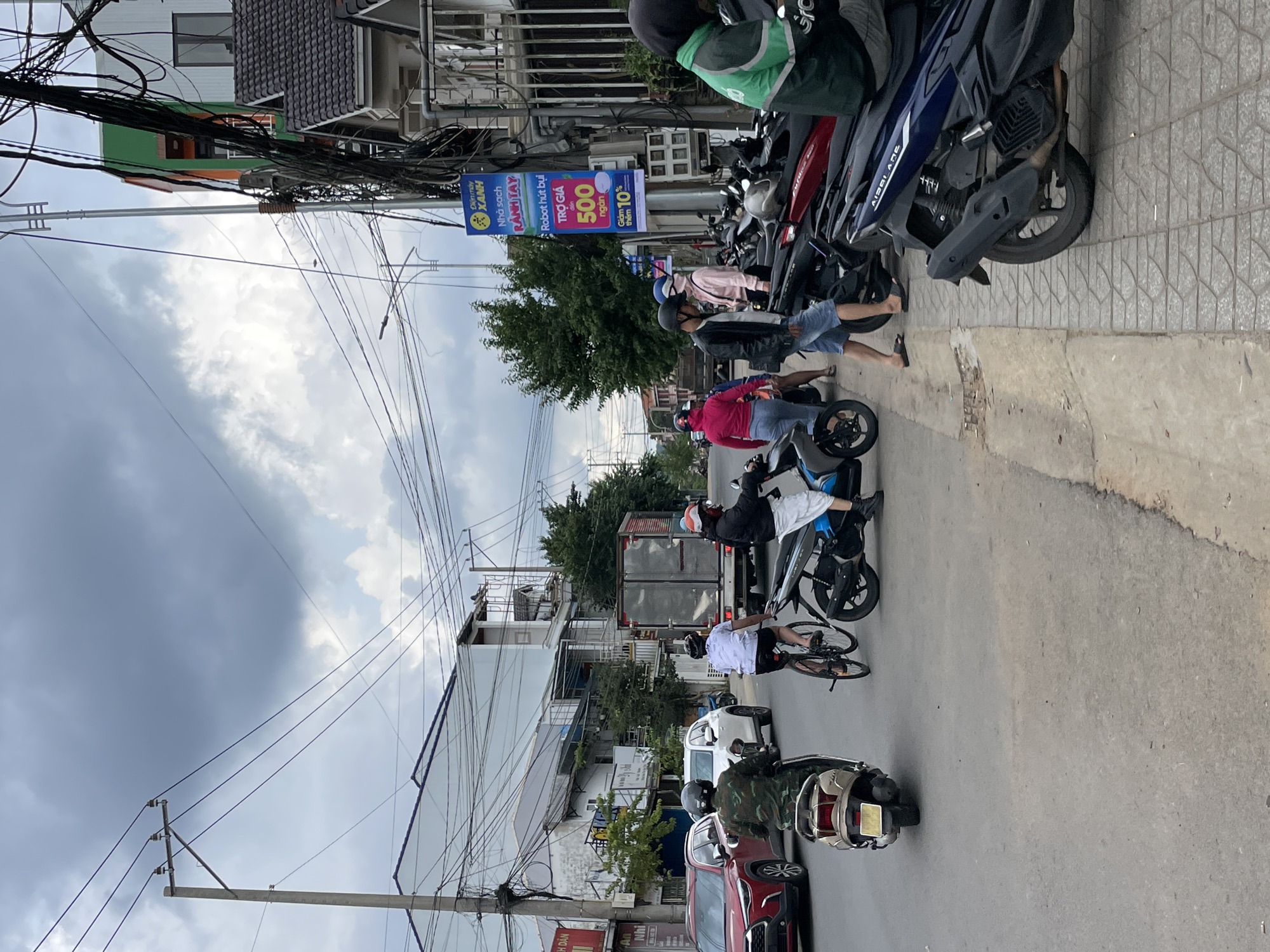 The image depicts a street scene in Lâm Viên - Đà Lạt, Vietnam, under a cloudy sky. Several individuals are present, primarily on or around motorcycles and bicycles. In the foreground, a person in a green jacket is seated on a dark blue motorcycle. To their left, a person wearing a black jacket and blue shorts is standing on the sidewalk next to a row of parked motorcycles. Further to the left, a person in a pink shirt and a person in a black jacket with a white helmet are standing.
On the street, a person is riding a bicycle, wearing a white t-shirt and black shorts. Another person on a scooter, dressed in a camouflage-patterned jacket, is also on the street. A red car is visible to the far left, with a white SUV behind it. A large delivery truck is in the background. The environment includes a sidewalk paved with patterned tiles, a street, buildings lining the road, numerous power lines suspended overhead, and trees. A blue and yellow sign with Vietnamese text is affixed to a pole on the right side of the street.