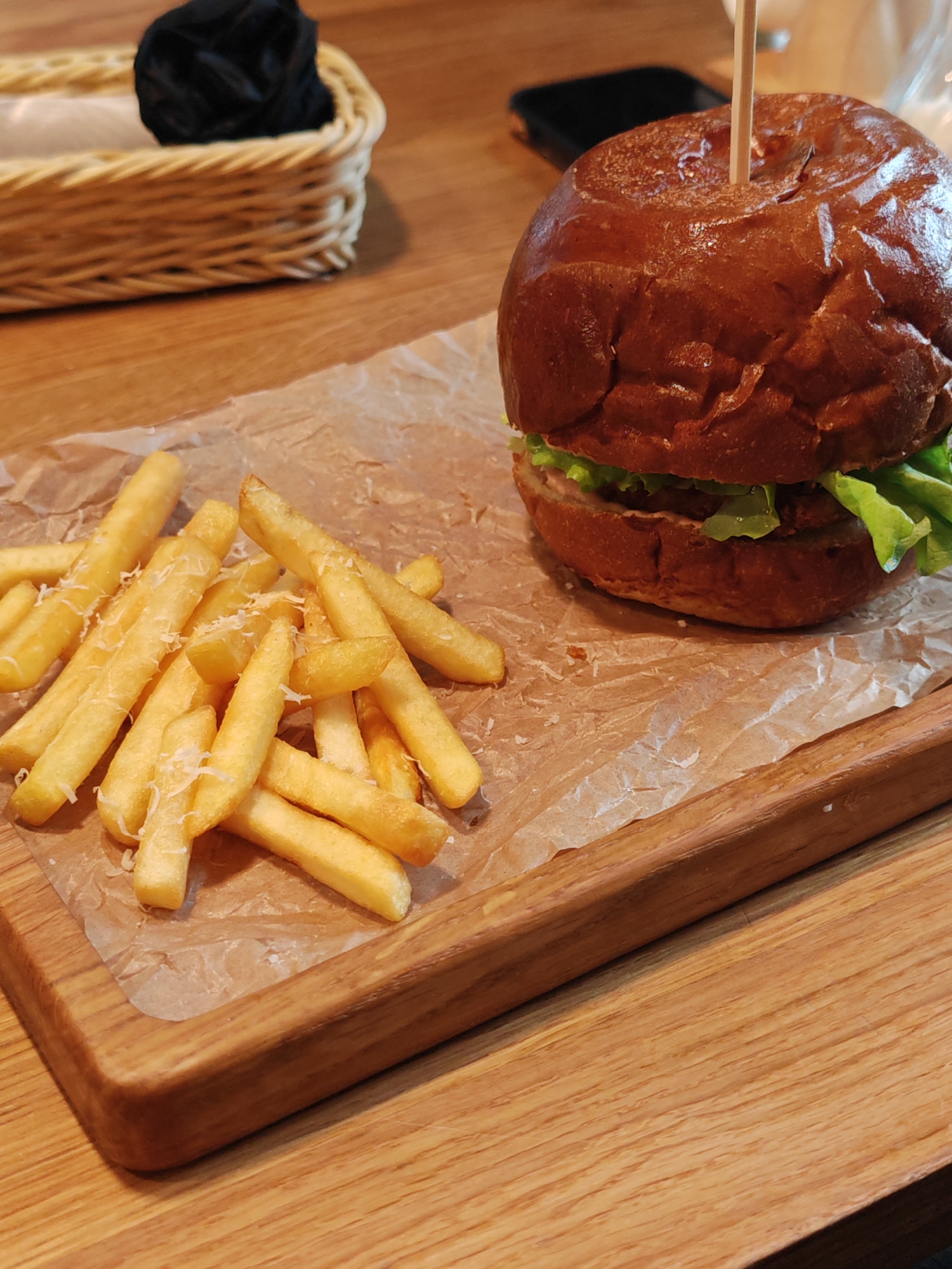 On a wooden table, presumably in a restaurant in Hrodna, Belarus, sits a wooden cutting board holding a burger and fries. The burger, secured with a wooden skewer, has a glossy brown bun, lettuce, and what appears to be a meat patty. Adjacent to the burger are a pile of golden fries sprinkled with grated cheese. In the background, a woven basket holding dark napkins and a black smartphone add context to the setting. The scene is a close-up shot, focused on the food.
