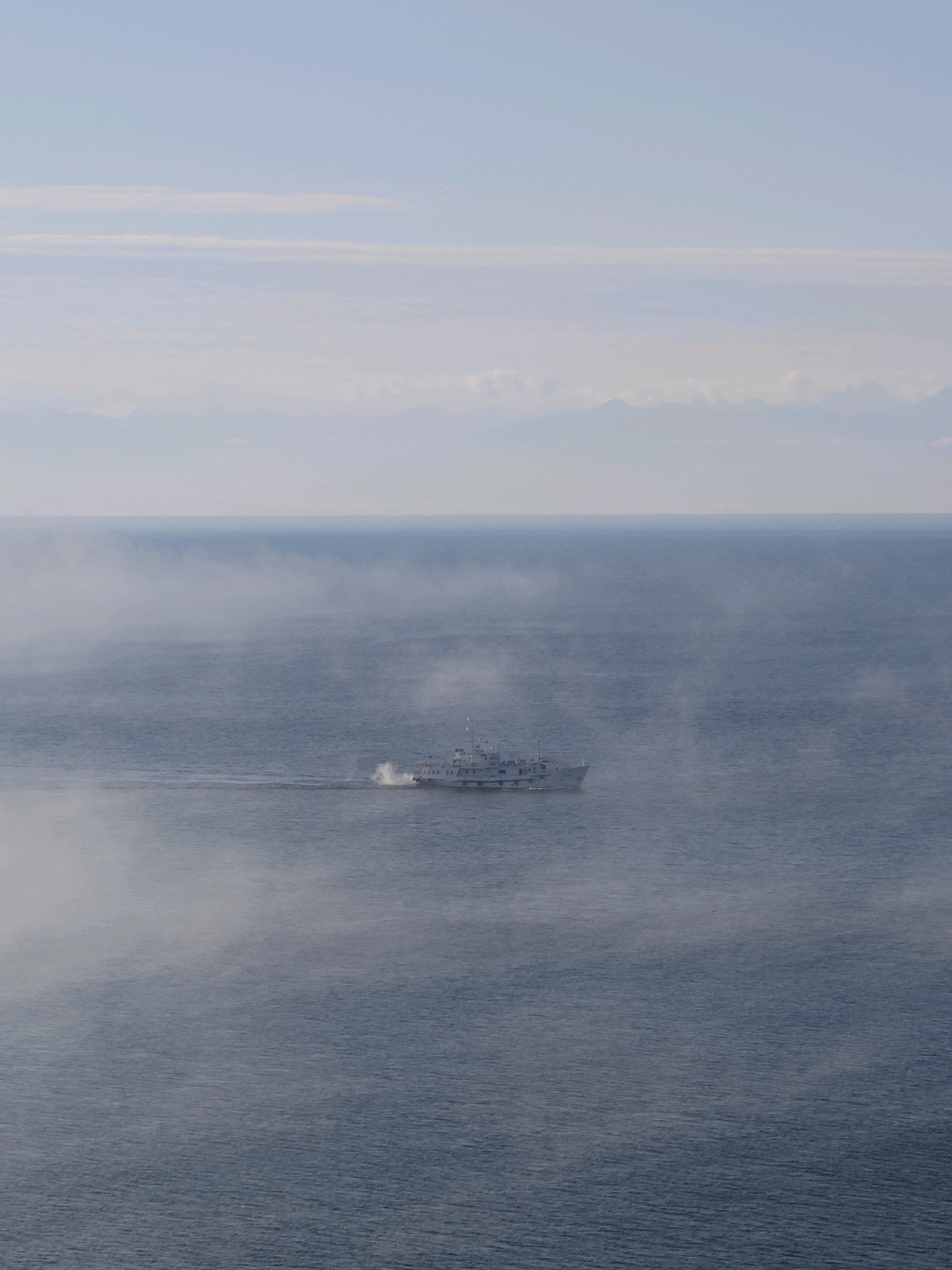 An aerial, slightly elevated shot captures a large body of water, likely Lake Baikal, Russia. The water is a deep blue-grey, with subtle ripples indicating calm conditions. A white passenger boat with multiple decks sails across the water from left to right, leaving a faint wake behind it. The boat is centrally located in the frame, near the horizon line. A thin layer of mist hovers close to the water's surface, partially obscuring the boat and creating a hazy effect. In the background, the sky is a pale blue with wispy white clouds. Distant, hazy mountain ranges are visible along the horizon line, their peaks softly outlined against the sky. The overall atmosphere is serene and somewhat mysterious due to the mist.