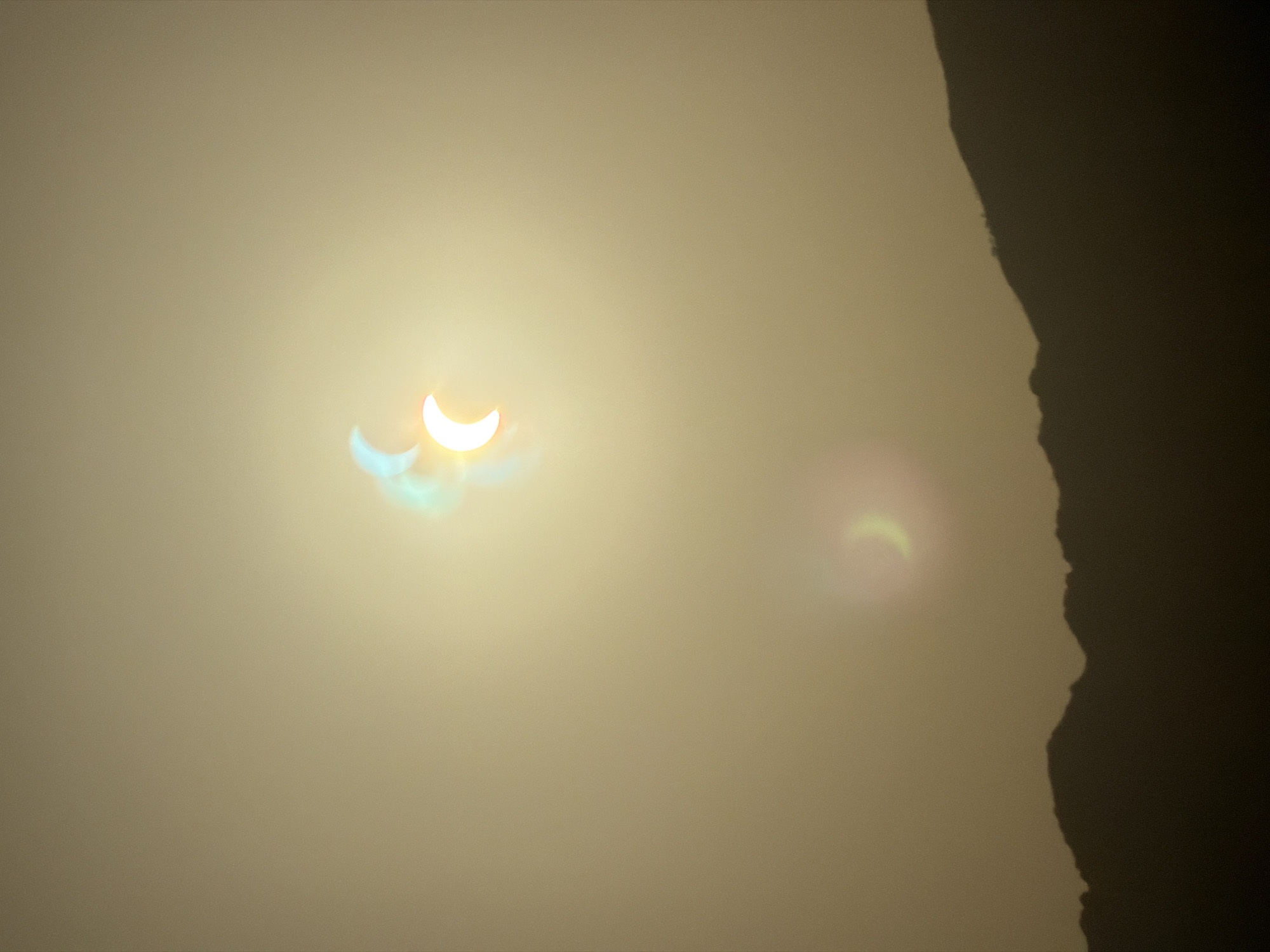 The image depicts a partial solar eclipse over the landscape of Resinera-voladilla, Spain. The sky is a hazy, yellowish-brown, obscuring any clear view of distant objects. Two crescent shapes, representing the eclipsed sun, are visible in the sky. The larger, brighter crescent is positioned towards the top center, emitting a bright white light with bluish and yellow flares around its edges. Below and to the left of this, a fainter, dimmer crescent is visible, appearing more yellow-green. In the foreground, the dark silhouette of a mountain range stretches across the bottom of the frame, its jagged peaks sharply defined against the muted sky. There are no people or identifiable structures visible in the image. The overall impression is one of a natural phenomenon observed in a remote or hazy environment.